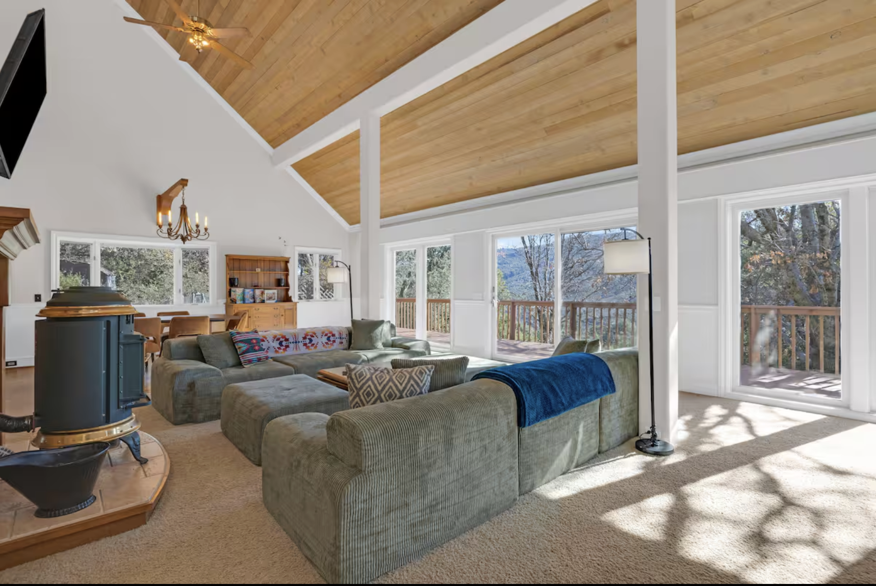 Warm, open living room at retreat venue with large windows, vaulted wooden ceiling, upholstered sectional sofa with throw pillows, a floor lamp, and a balcony view of trees and mountains outside.