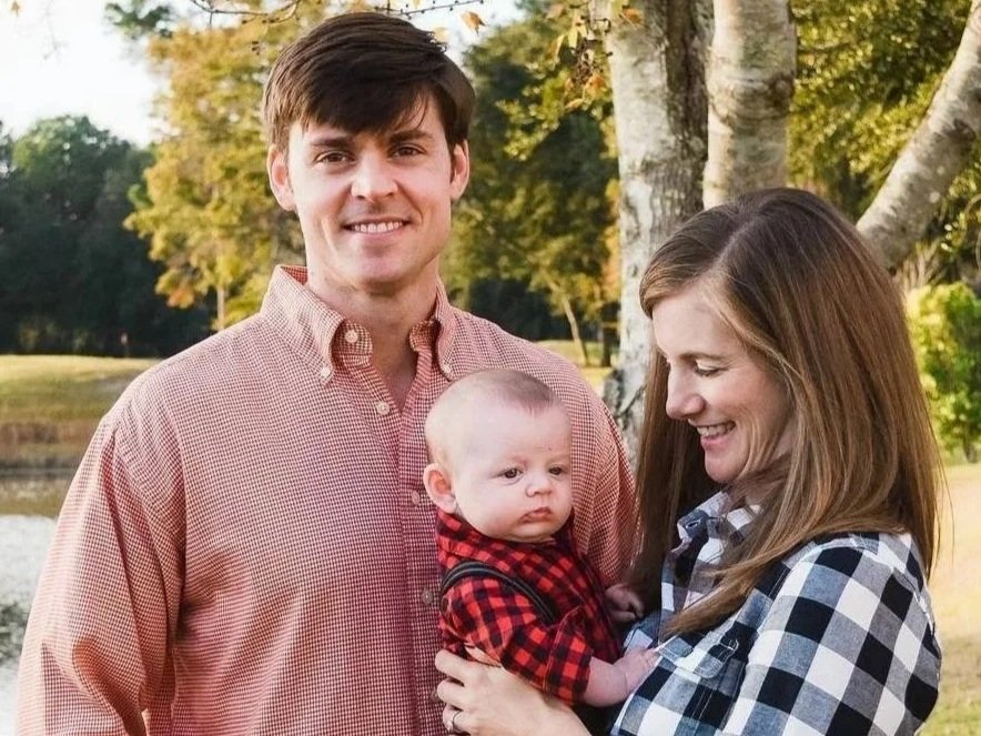 A family of three outdoors near a tree and a lake, with the mother holding a baby boy and the father standing beside them, all smiling.