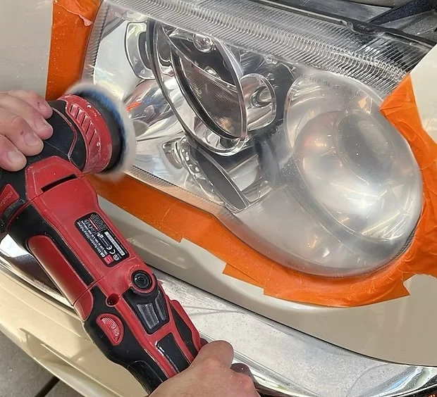 Person polishing a vehicle's headlight with a red electric rotary tool.