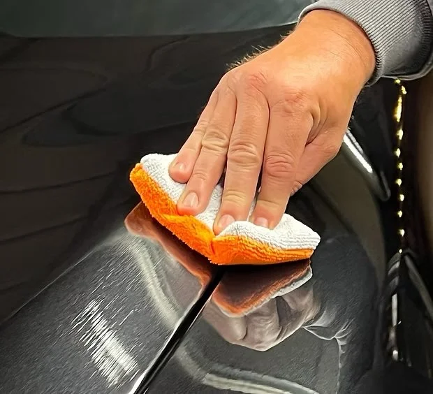 A person using a white and orange microfiber cloth to polish or clean a dark metallic surface, possibly a car.