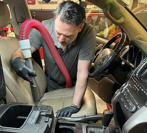 A local cape cod man in a gray shirt and black gloves vacuuming the interior of a car with a red vacuum hose.