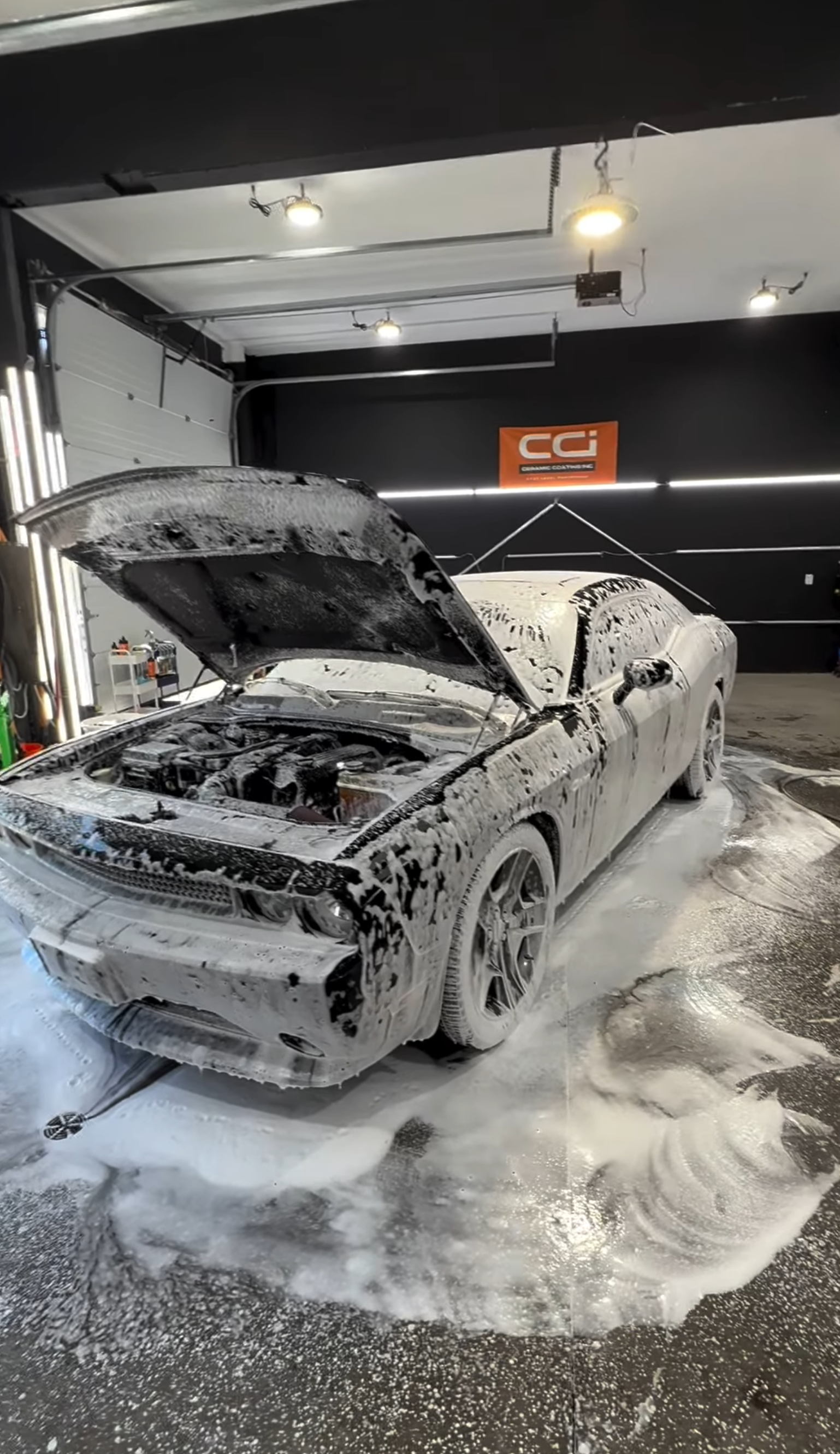 A silver sports car covered in soap suds, with its hood open, inside a garage with black walls and a sign that says 'CCI' in the background.