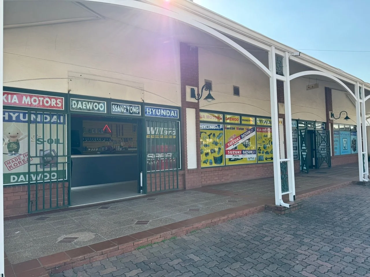 Car repair shop with signs for Hyundai, Daewoo, Ssang Yong, Kia Motors, Suzuki, Datsun, and parts. The shop has an open entrance secured by metal gates, informational posters, and a brick sidewalk covered by a white canopy.