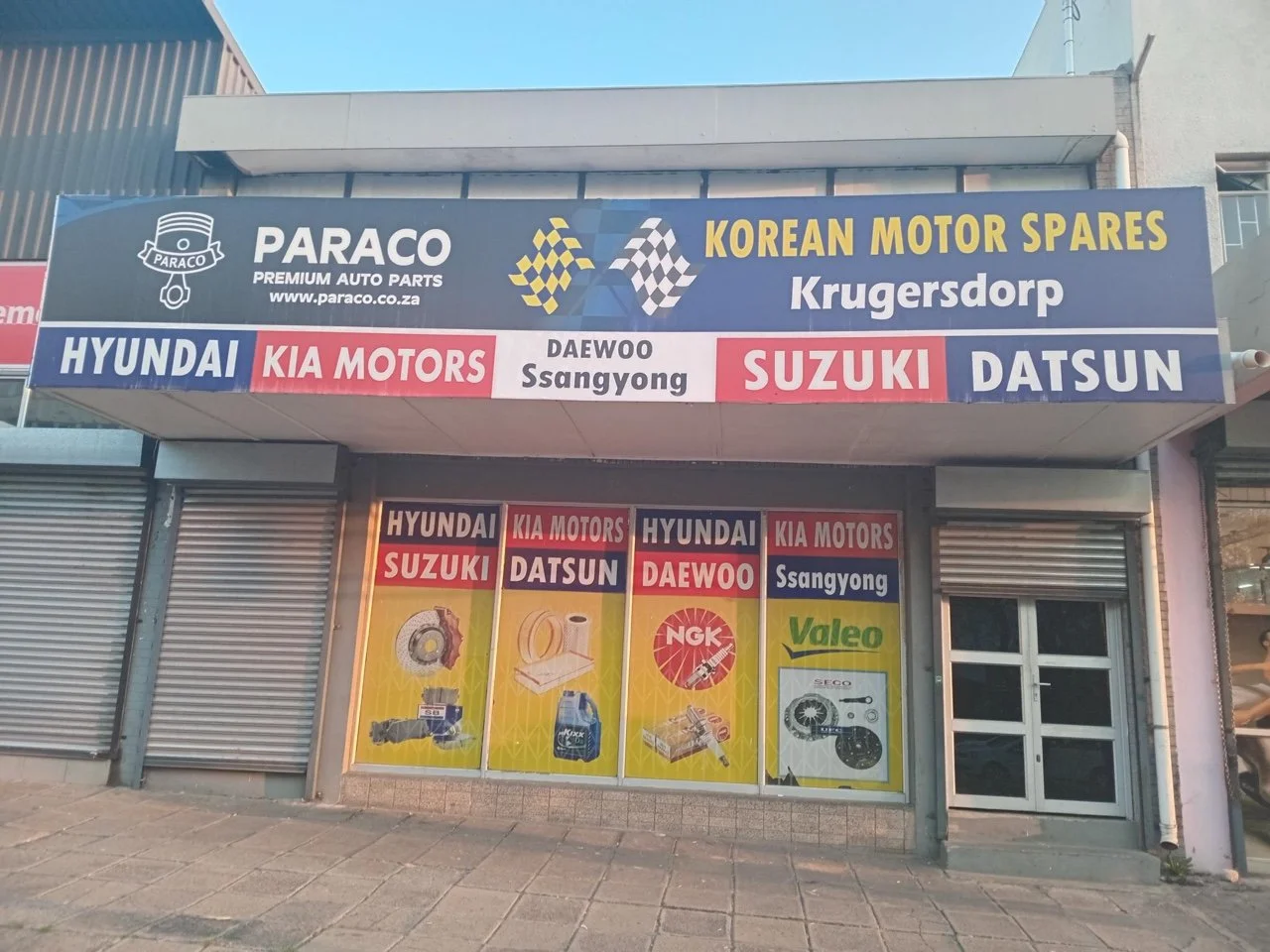 Signboard of a Korean motor spares shop. The sign lists brands like Hyundai, Kia Motors, Suzuki, Datsun, Daewoo Ssangyong, with images of auto parts below, inside a commercial building with closed metal shutters.