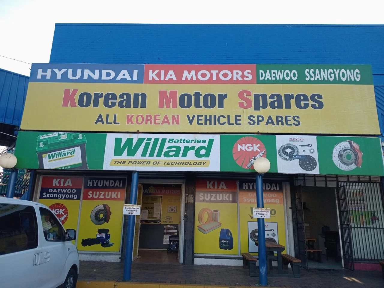 Korean motor spare parts store with signs for Hyundai, Kia, Daewoo, Ssangyong, and Suzuki, and advertisements for car batteries, NGK spark plugs, and brakes.