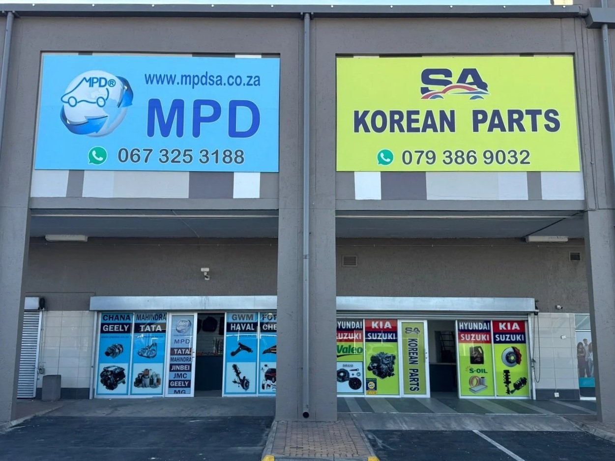 Exterior view of a building with two large signs on the upper level. The left sign is light blue and promotes MPD auto parts with a website, phone number, and WhatsApp icon. The right sign is yellow-green and promotes Korean car parts with a WhatsApp icon and phone number. The lower level has storefront windows with images and logos of car brands and auto parts, including Hyundai, Kia, Suzuki, and others.