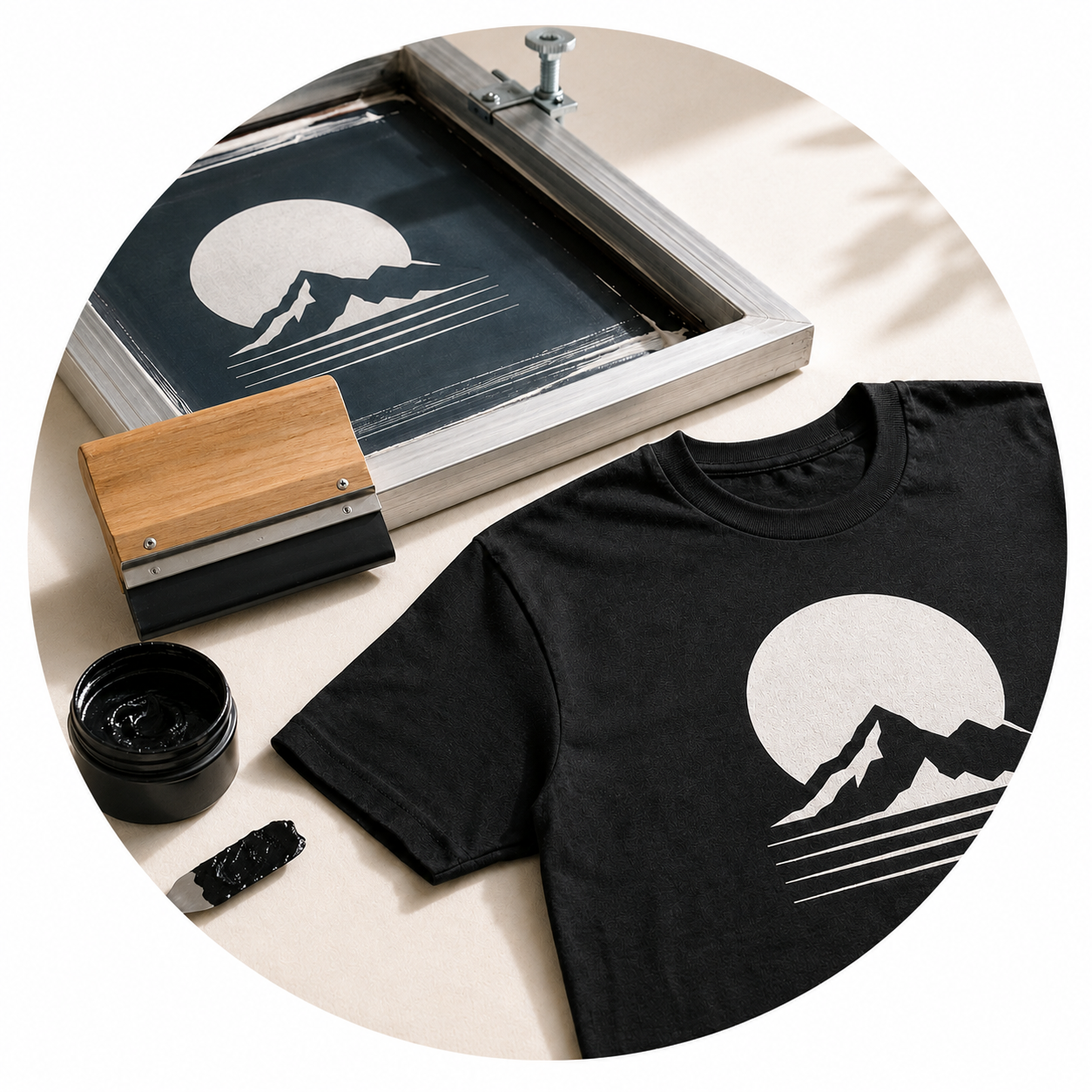 A black T-shirt with a mountain and sun graphic, a screen printing frame, a container of black ink, and a squeegee with black ink on it, all set up for screen printing.
