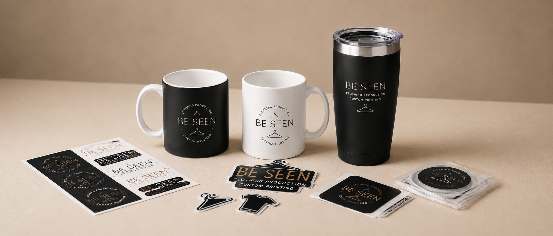 Two ceramic mugs, one black and one white, with the logo "BE SEEN" and text related to clothing production and custom printing. A black travel tumbler with the same logo and text is positioned to the right. Various stickers, tags, and a clear plastic case containing additional stickers are arranged on a beige surface in front of the mugs.