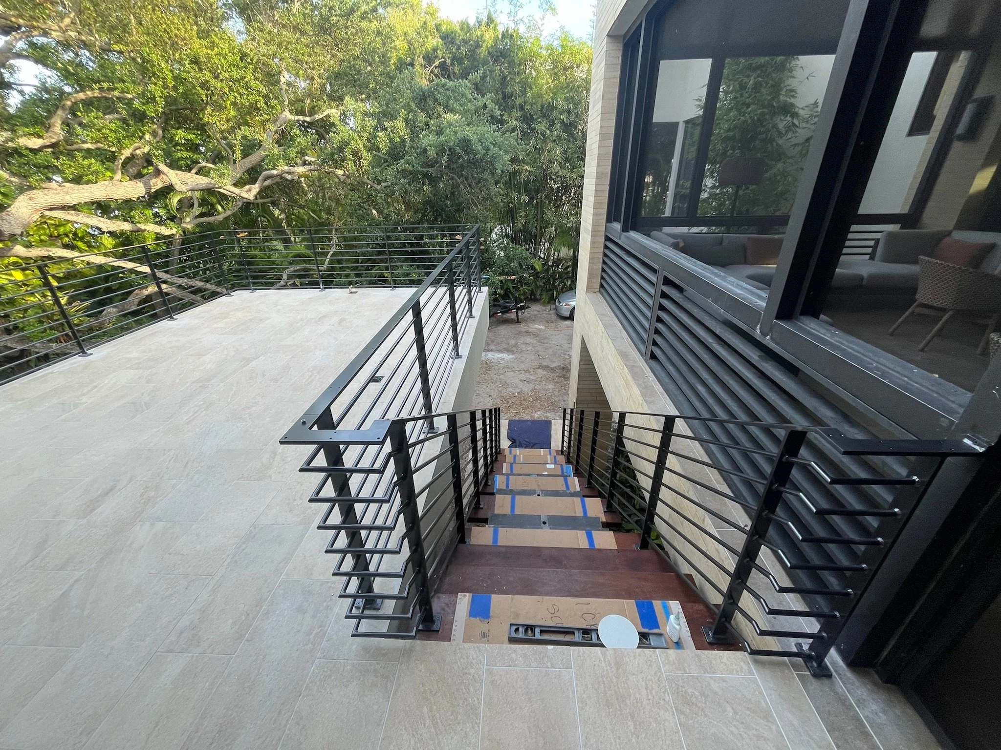 A building balcony with black metal railings, adjacent to a large window reflecting outdoor greenery. The balcony surface has beige tiles, and there are cardboard boxes with markings placed on the floor near the window. Trees and outdoor foliage are 