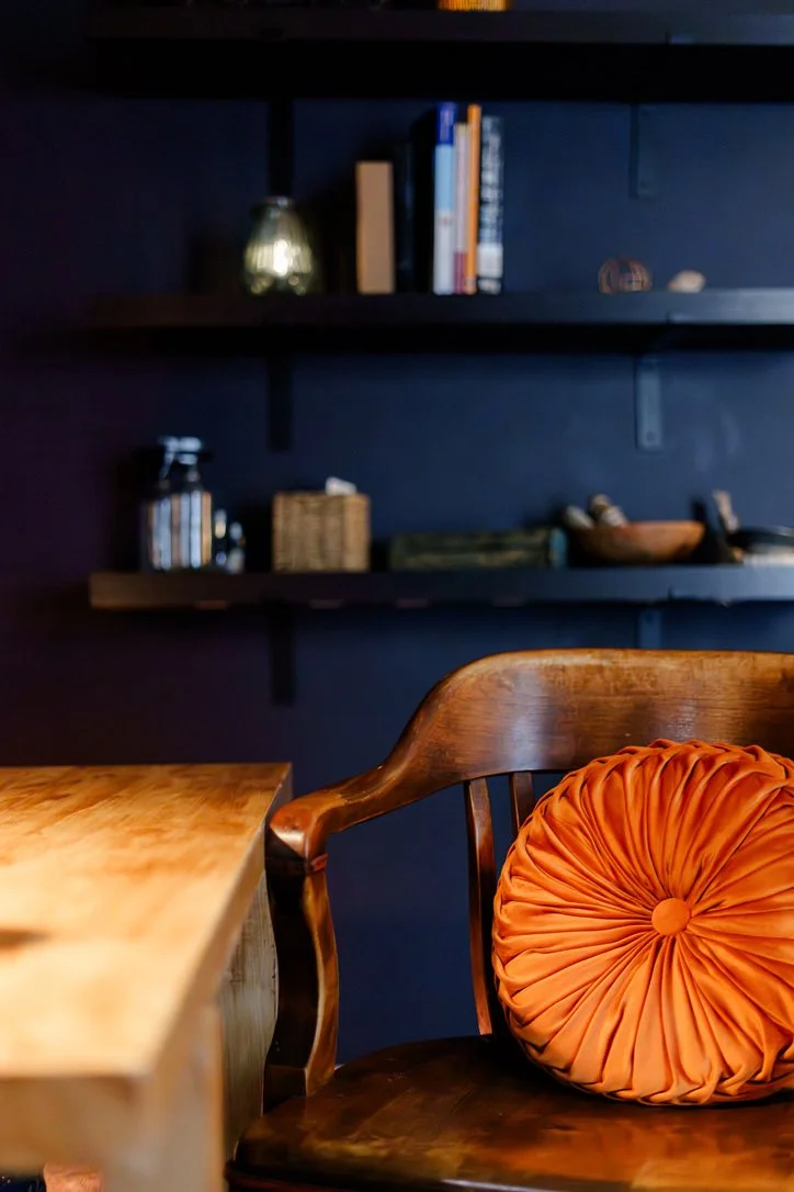 A wooden chair with an orange round cushion in a room with dark blue walls and floating black shelves holding books, decorative objects, and a small lamp.
