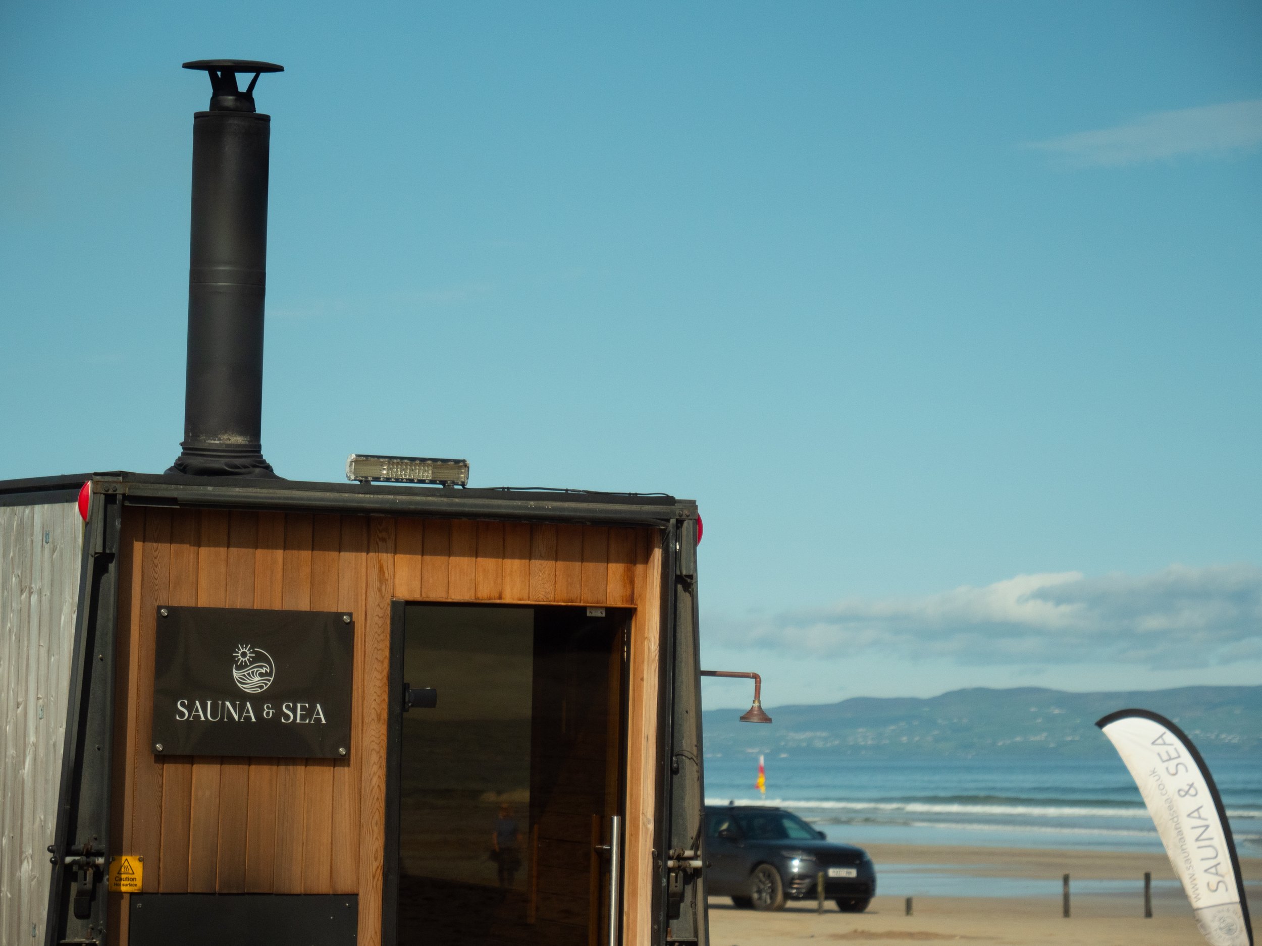 Wooden structure with a sign that says "Sauna & Sea" and a black chimney pipe on top, located on a beach with a view of the sea and mountains in the distance.
