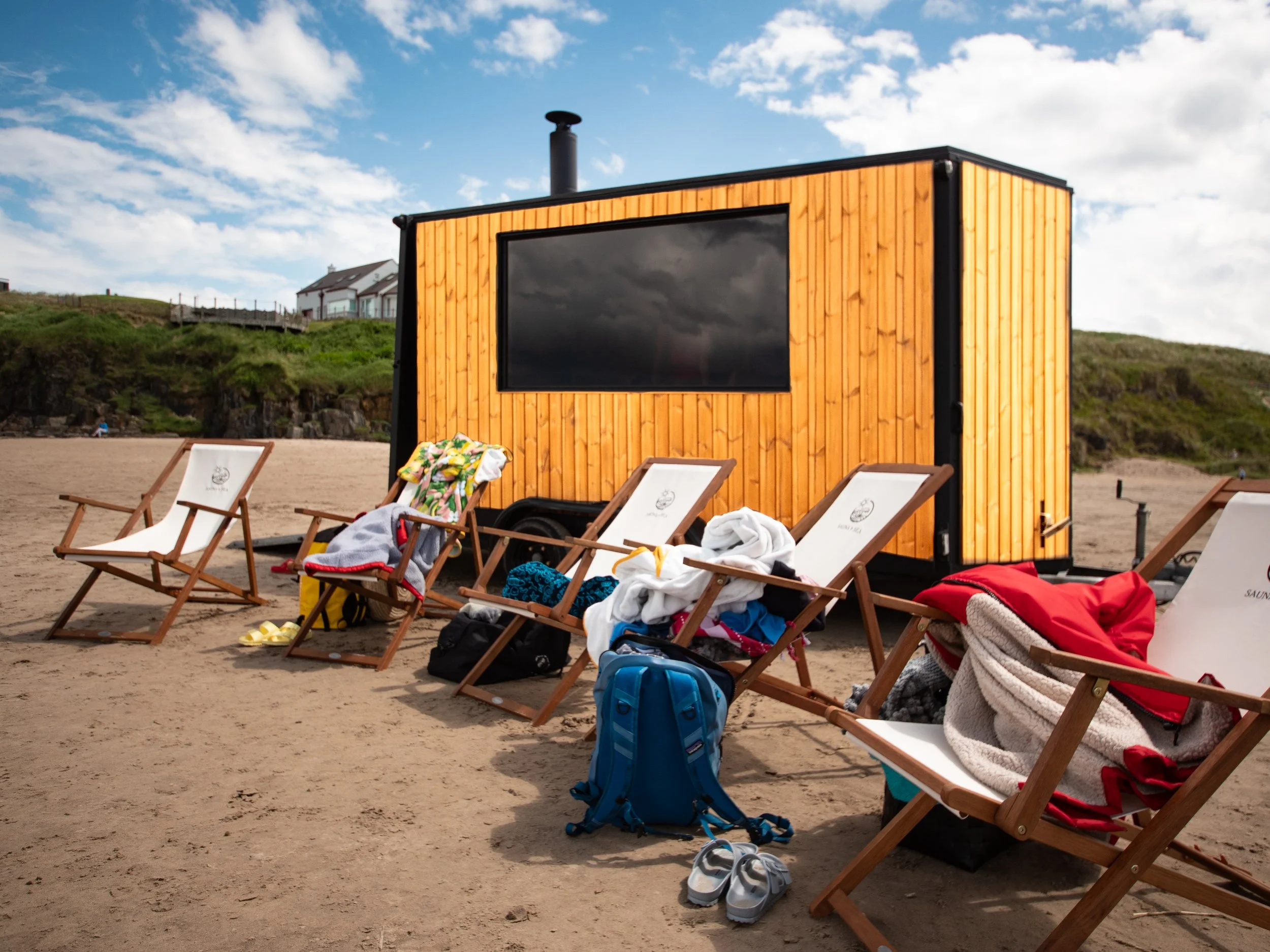 Beach scene on Portstewart strand, a national trust site, with wooden lounge chairs, some covered with clothes and towels, and backpacks on the sand. A mobile sauna is set up behind and people appear to be in it.