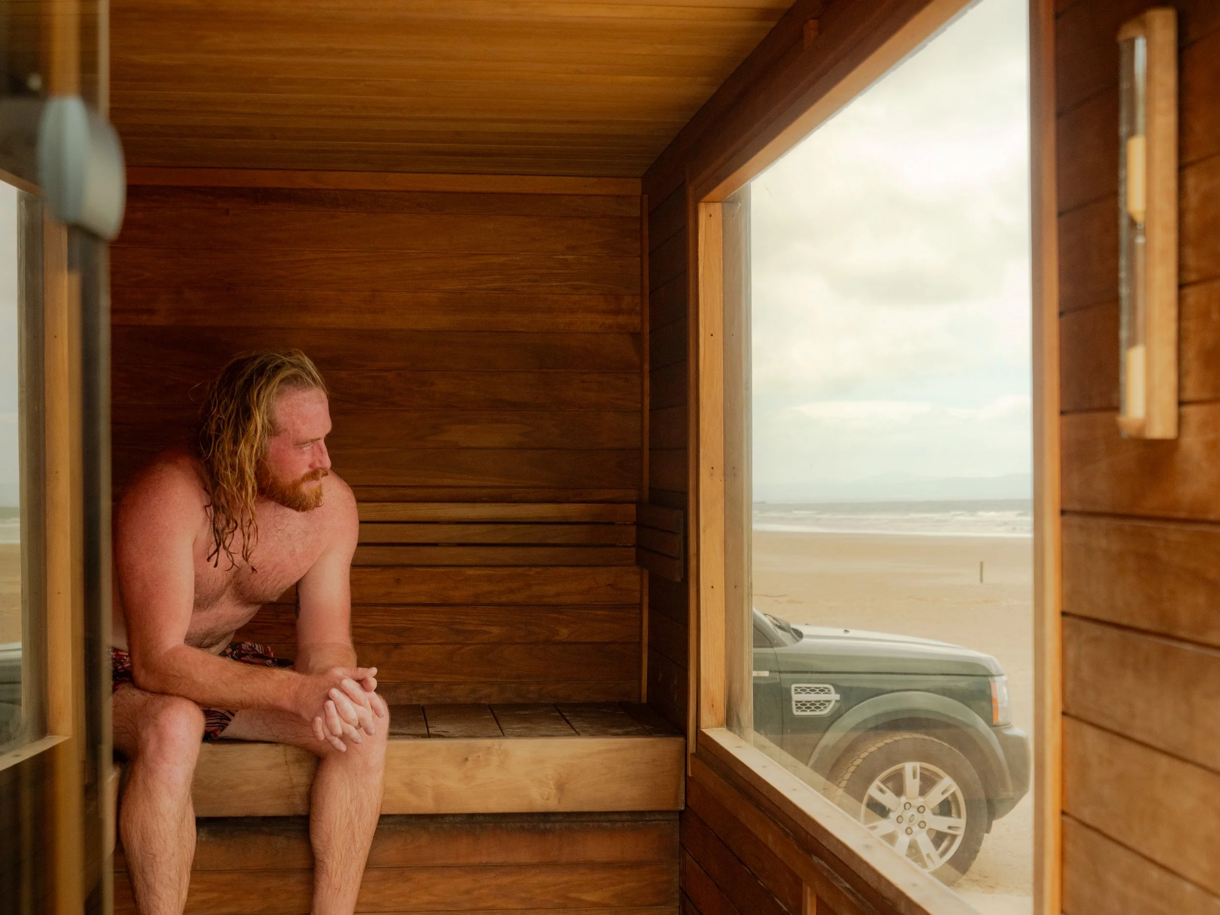 A man with long, curly red hair and a beard sits in a wooden sauna, looking down with his hands clasped. Outside the sauna windows, a washing machine and a cloudy sky are visible.