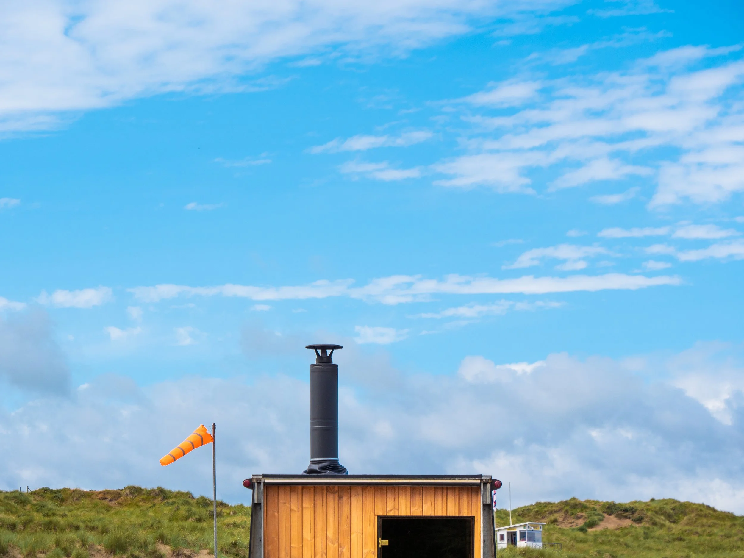 Image of a weather station with a wind cone next to a tall black vent pipe against a partly cloudy sky. Small building with a wooden facade is visible at the bottom of the image.