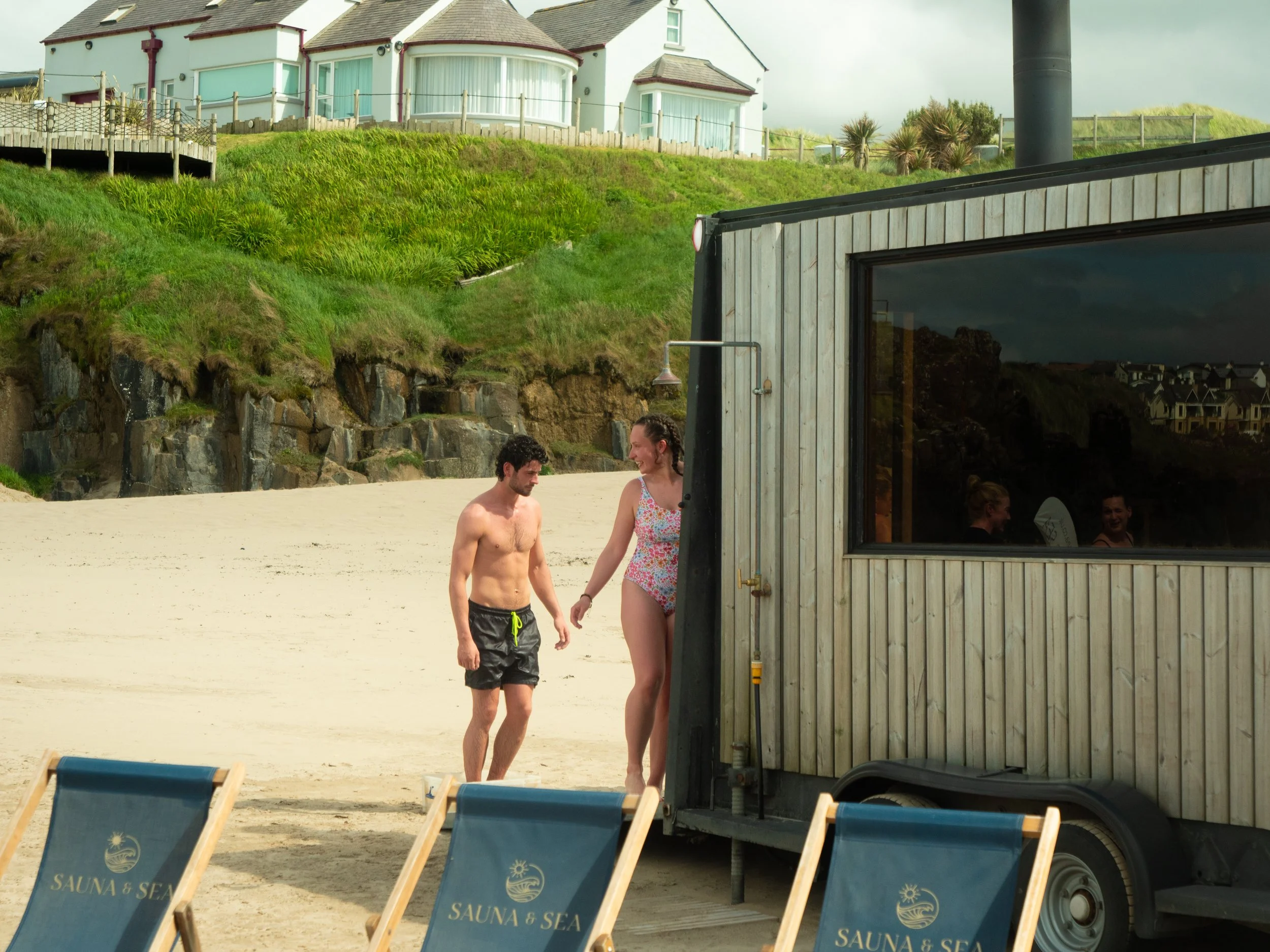 People walking on a sandy beach near a small wooden building with a large window. The beach has lounge chairs and a green grassy hill with houses on top in the background.