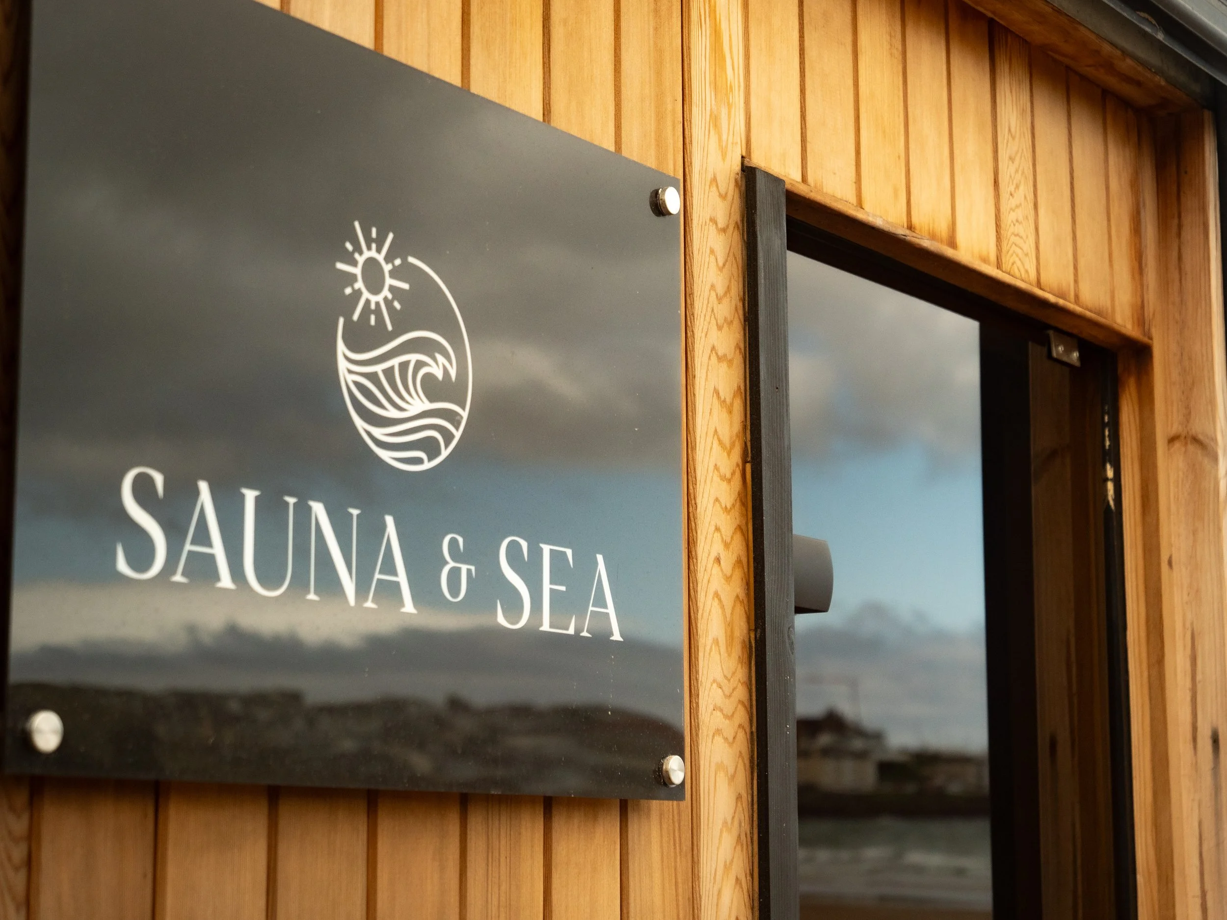 Close-up of a sign reading 'SAUNA & SEA' mounted on a wooden wall with horizontal planks, with a black background and white lettering, including a logo of a sun and wave.