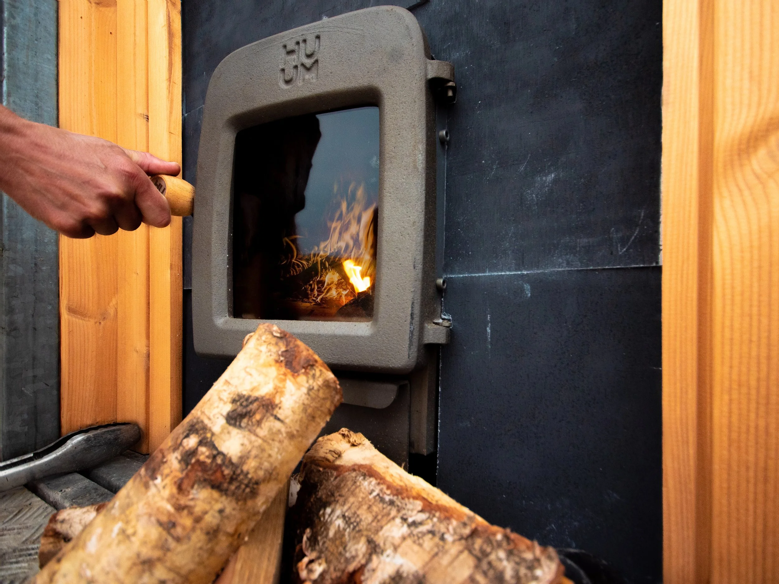 A hand holding a matchstick over a small indoor fireplace with logs inside, ready to ignite. The fireplace is set against a black wall with wooden paneling around it.