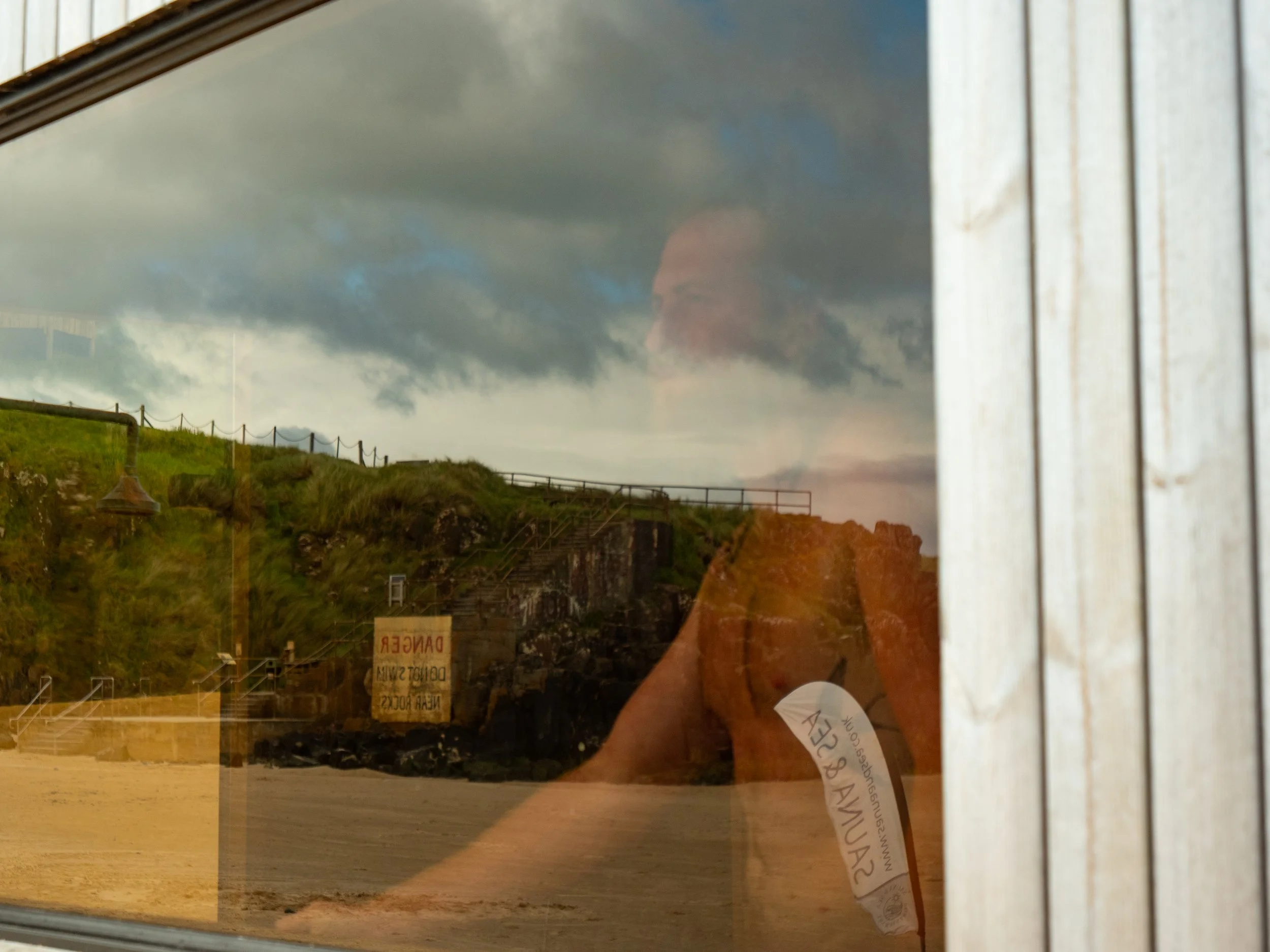 Reflections of a cloudy sky and a person with glasses in a window outside a building with a fence and a 'Danger' sign.