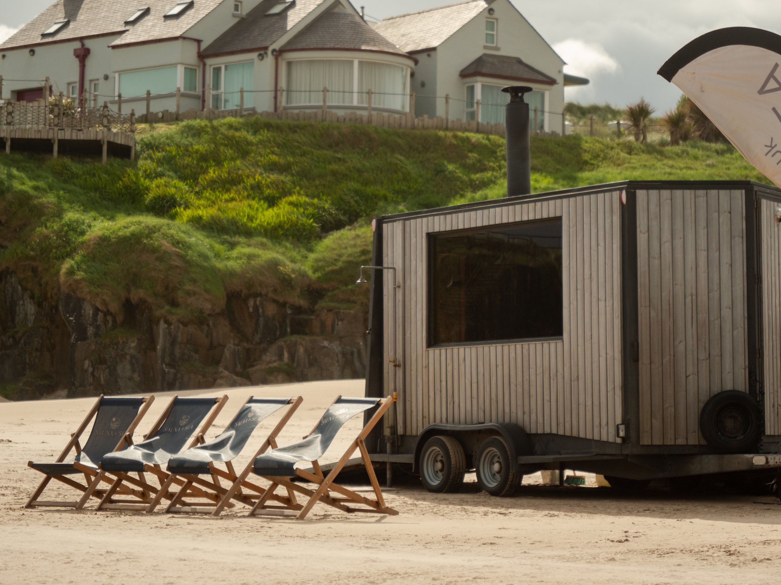 Beach scene with wooden deck chairs in a row, a food truck or trailer, and a house in the background on a grassy hill.
