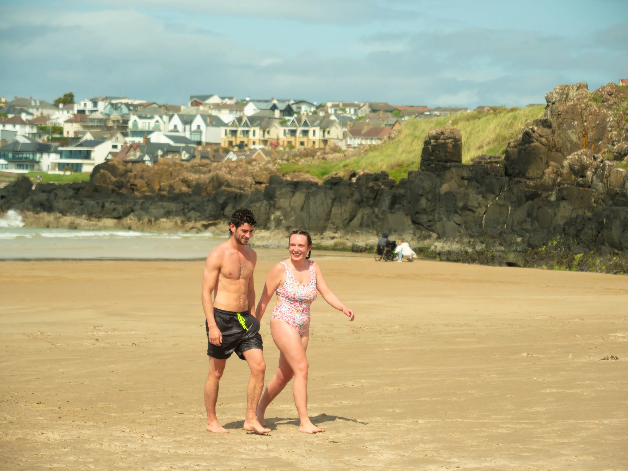 A man and a woman walk arm in arm on a national trust site portstewart strand, with rocks and a grassy hill in the background, and houses on the hill. They seem to be engaging in sea dipping activities.