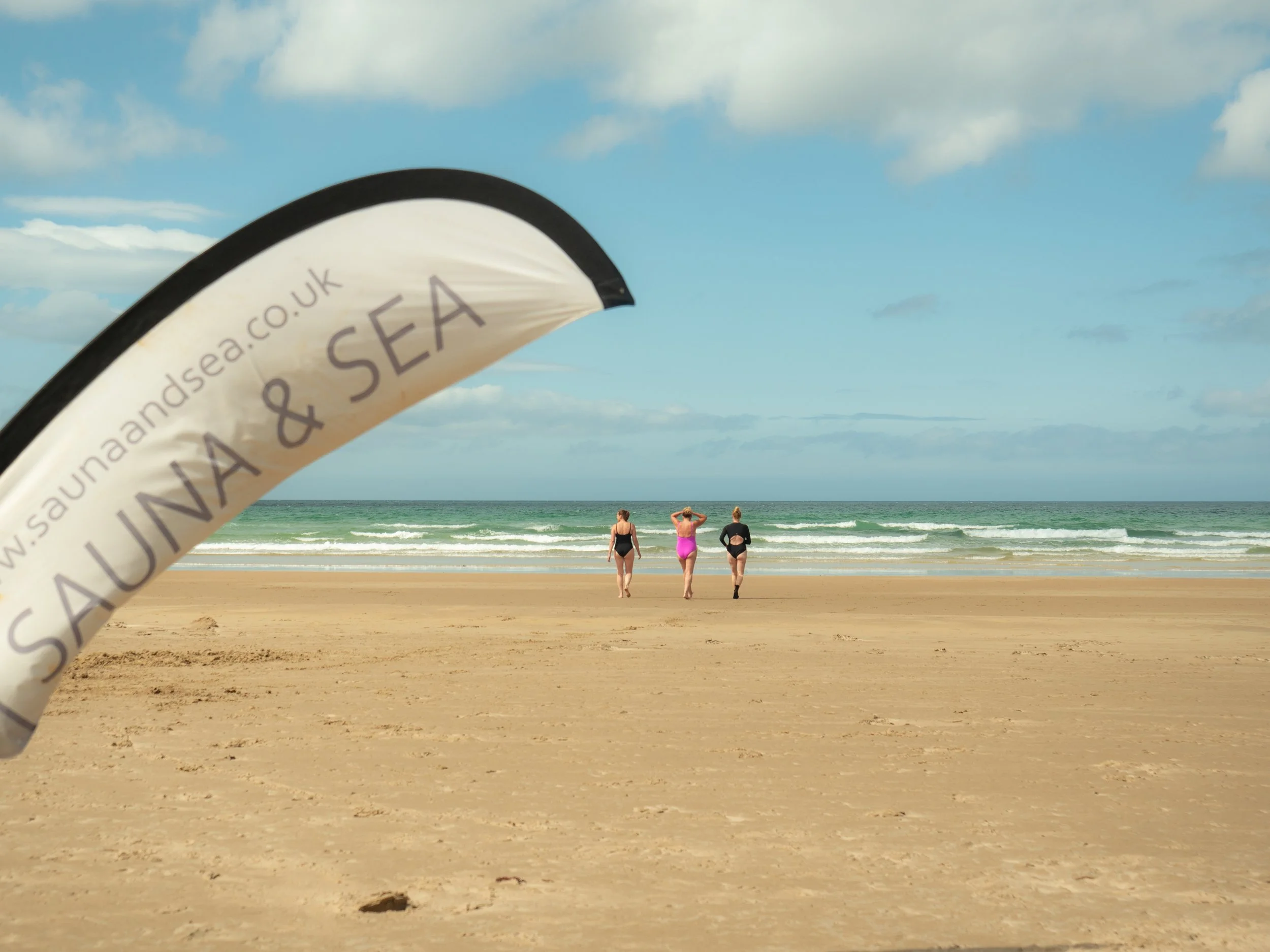 A beach scene with three women walking towards the ocean, a large flag in the foreground with text about surfing, and a partly cloudy sky.