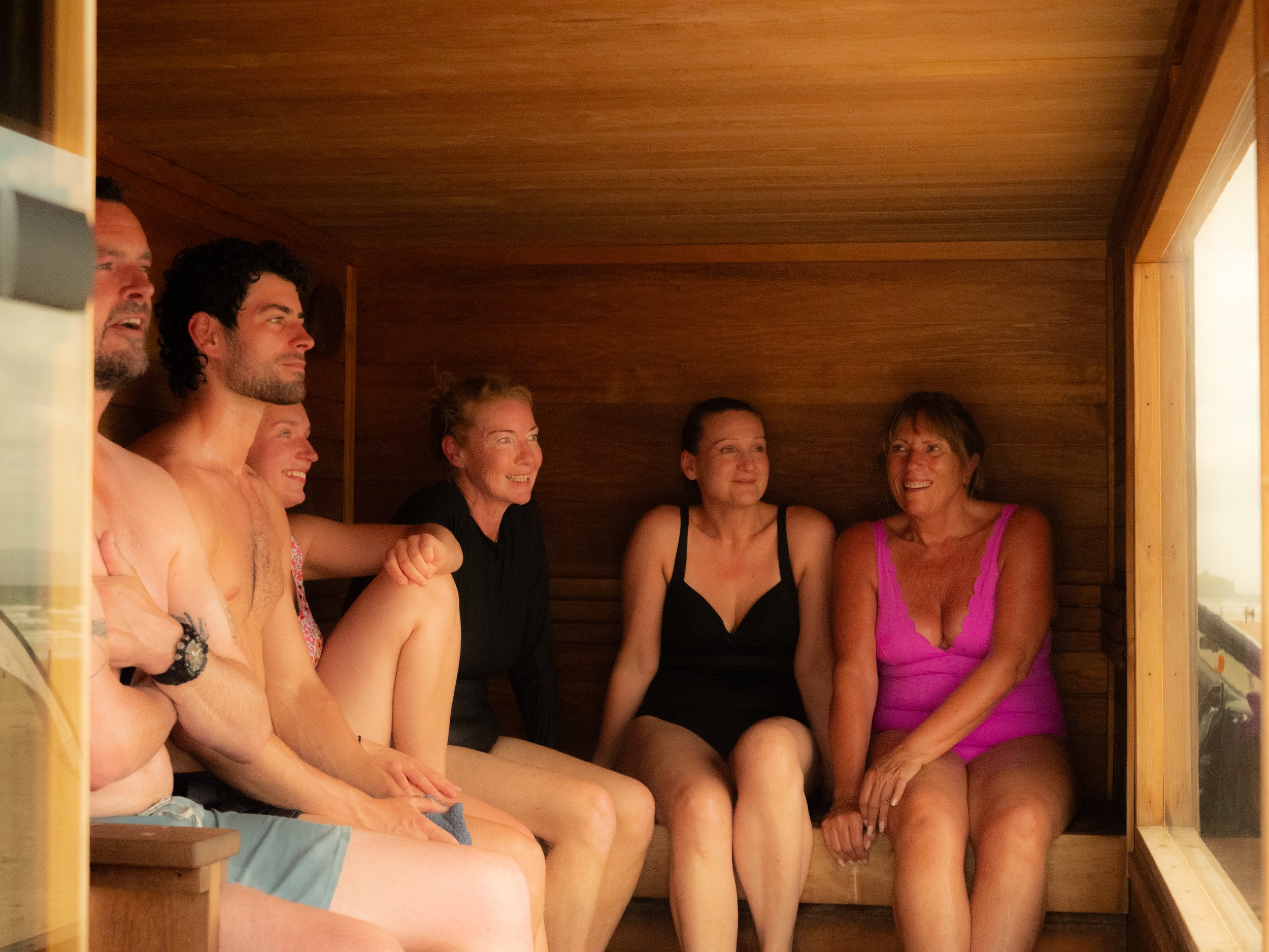 A group of seven people, five women and two men, sitting inside a wooden sauna, smiling and engaging in conversation at Sauna and Sea Portstewart.