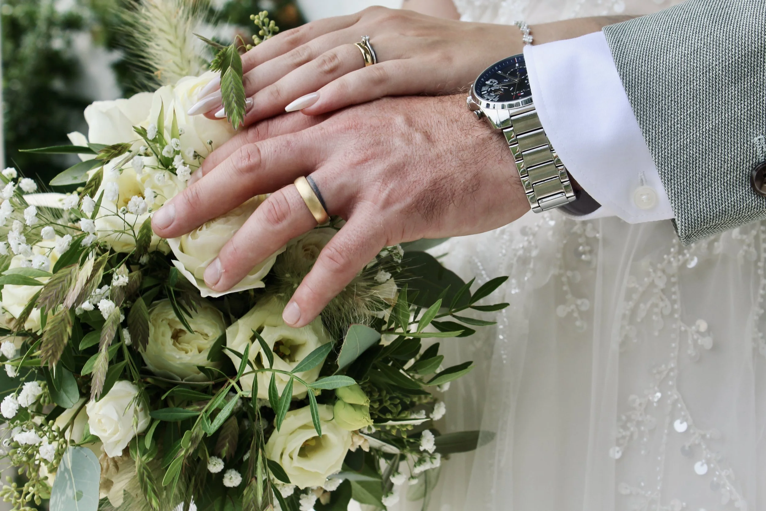 Nahaufnahme der Hände eines Paares mit Eheringen, einer Armbanduhr und einem weissen Blumenstrauss, im Hintergrund eine Hochzeitskleidung.