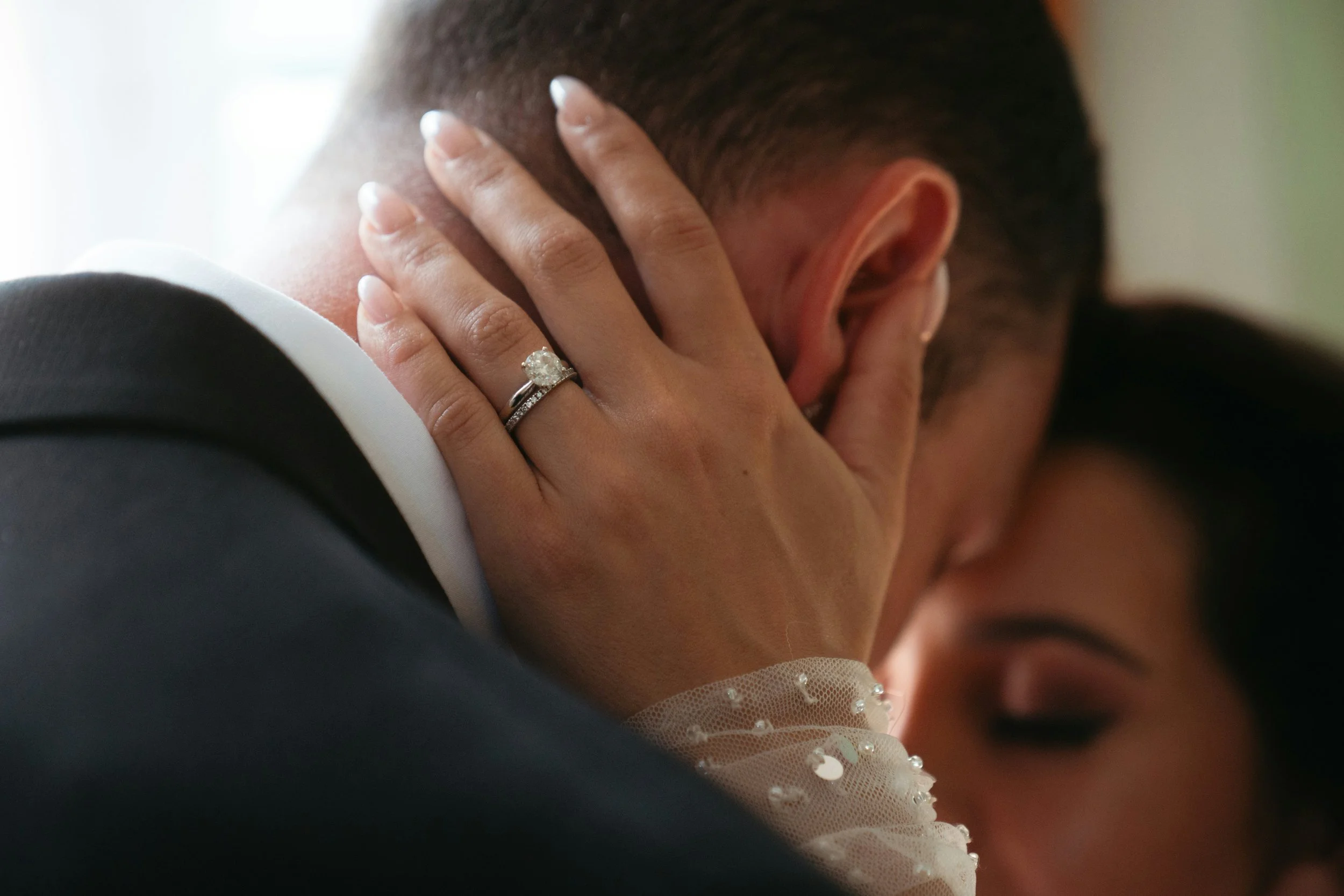 Nahaufnahme eines Paares bei einer Hochzeitszeremonie. Eine Frau trägt einen Ehering und ein elegantes Hochzeitskleid, während sie den Kopf ihres Bräutigams berührt, der eine dunkle Jacke trägt.