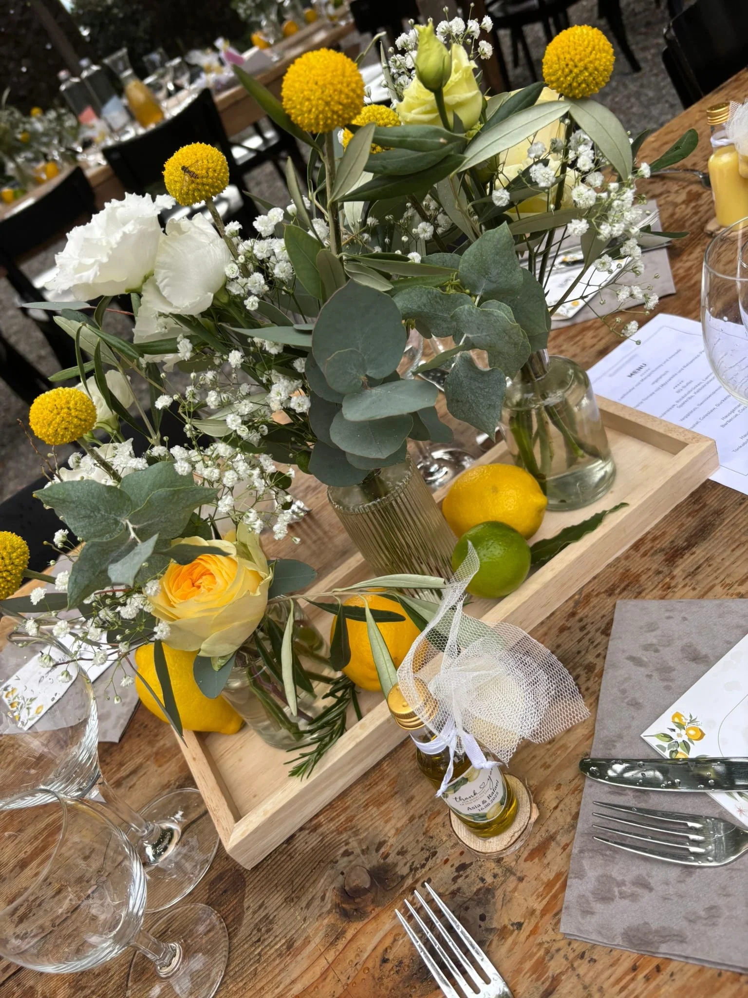 Blumenstrauss mit weissen Rosen, gelben Craspedia und kleinen weissen Blumen in einem Glasvasen auf einem Holztisch, daneben Zitronen und Limoncello-Flaschen, ein Namensschild und Besteck.