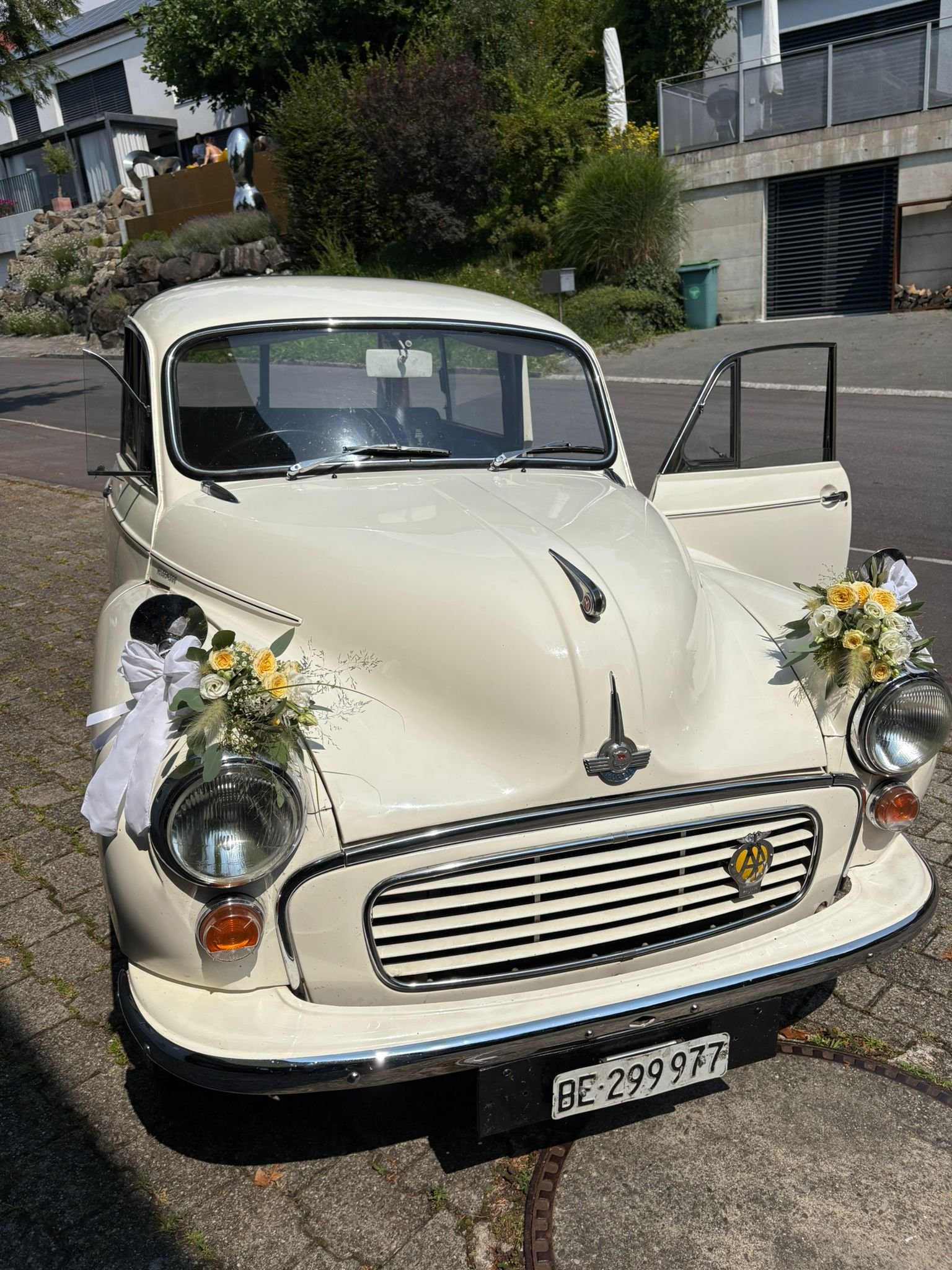 Ein weisses, vintage Auto, dekoriert mit Blumen für eine Hochzeit, parkend auf einer Strasse in einer modernen Wohngegend, bereit für den Hochzeitseinzug.