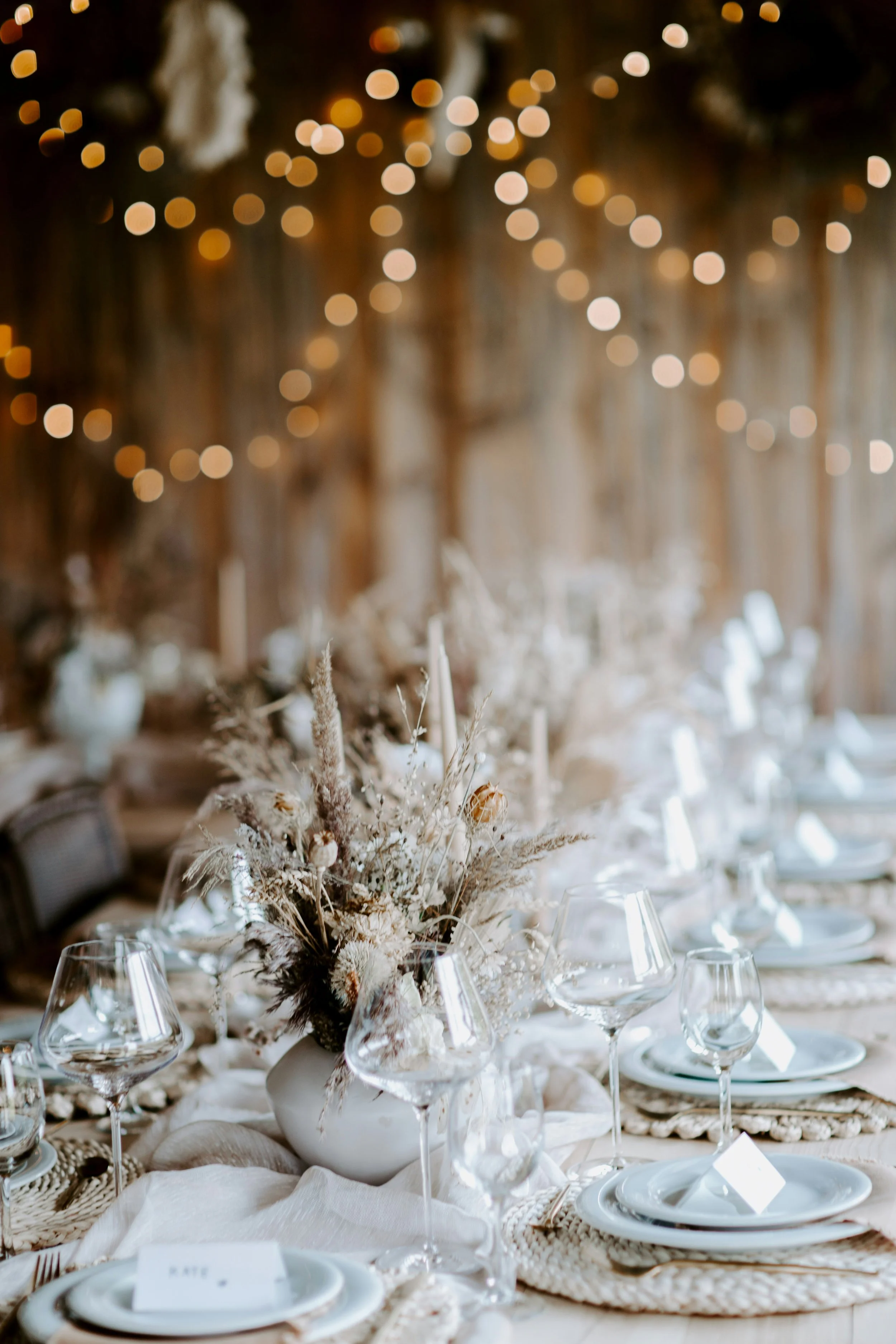 Eleganter Esstisch mit weisser Tischdecke, Blumenarrangement in Vase, Weingläsern, weissen Tellern und Platzkarten, im Hintergrund festliche Beleuchtung und ein rustikaler Holz-Hintergrund, liebevoll angerichtet.