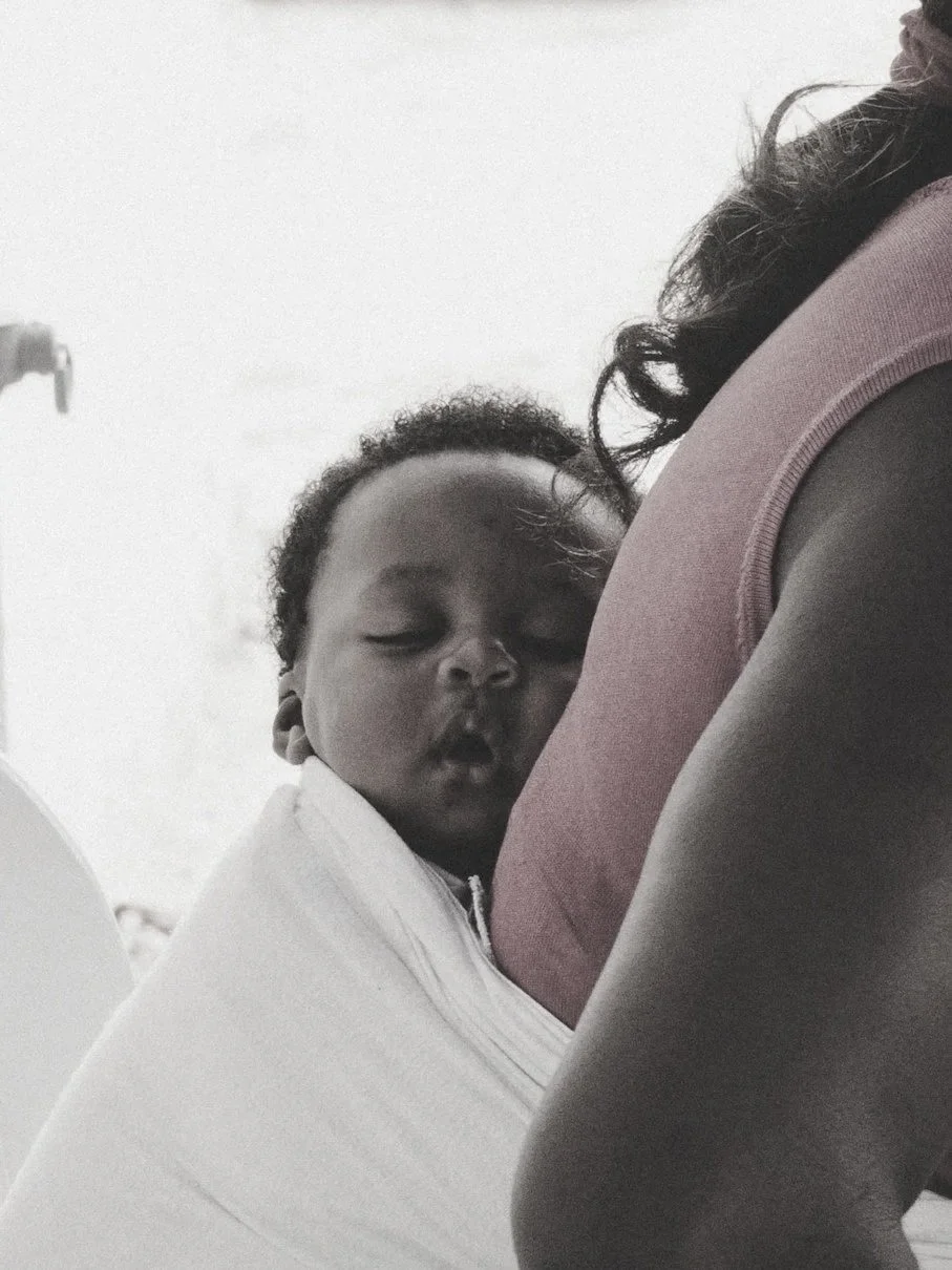 A young child is sleeping with their head resting against a woman's chest, who is wearing a pink top. The child appears peaceful and is holding onto the woman's side as she supports them.