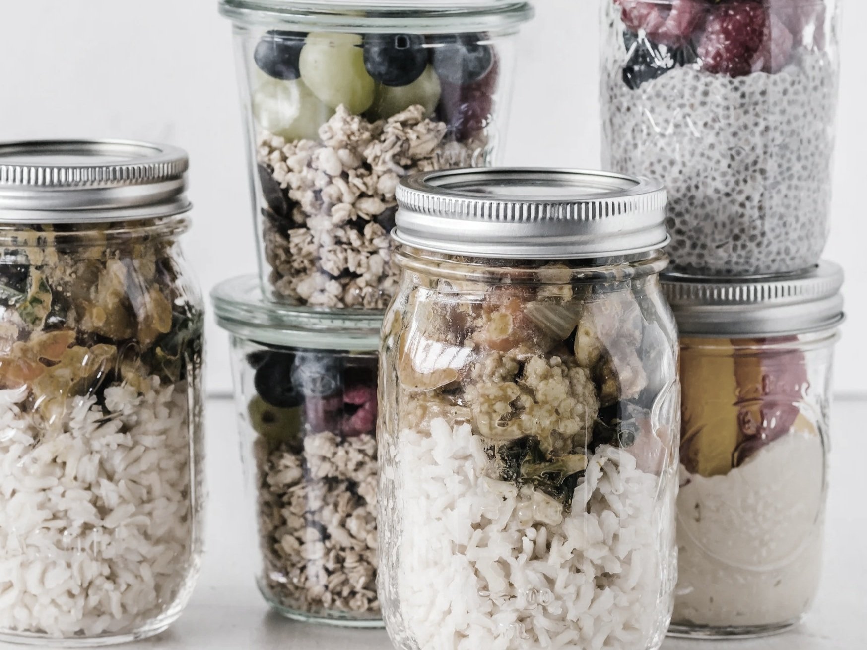 Multiple glass jars filled with various dried foods and snacks, including rice, granola, dried fruits, and seeds, arranged on a white surface.