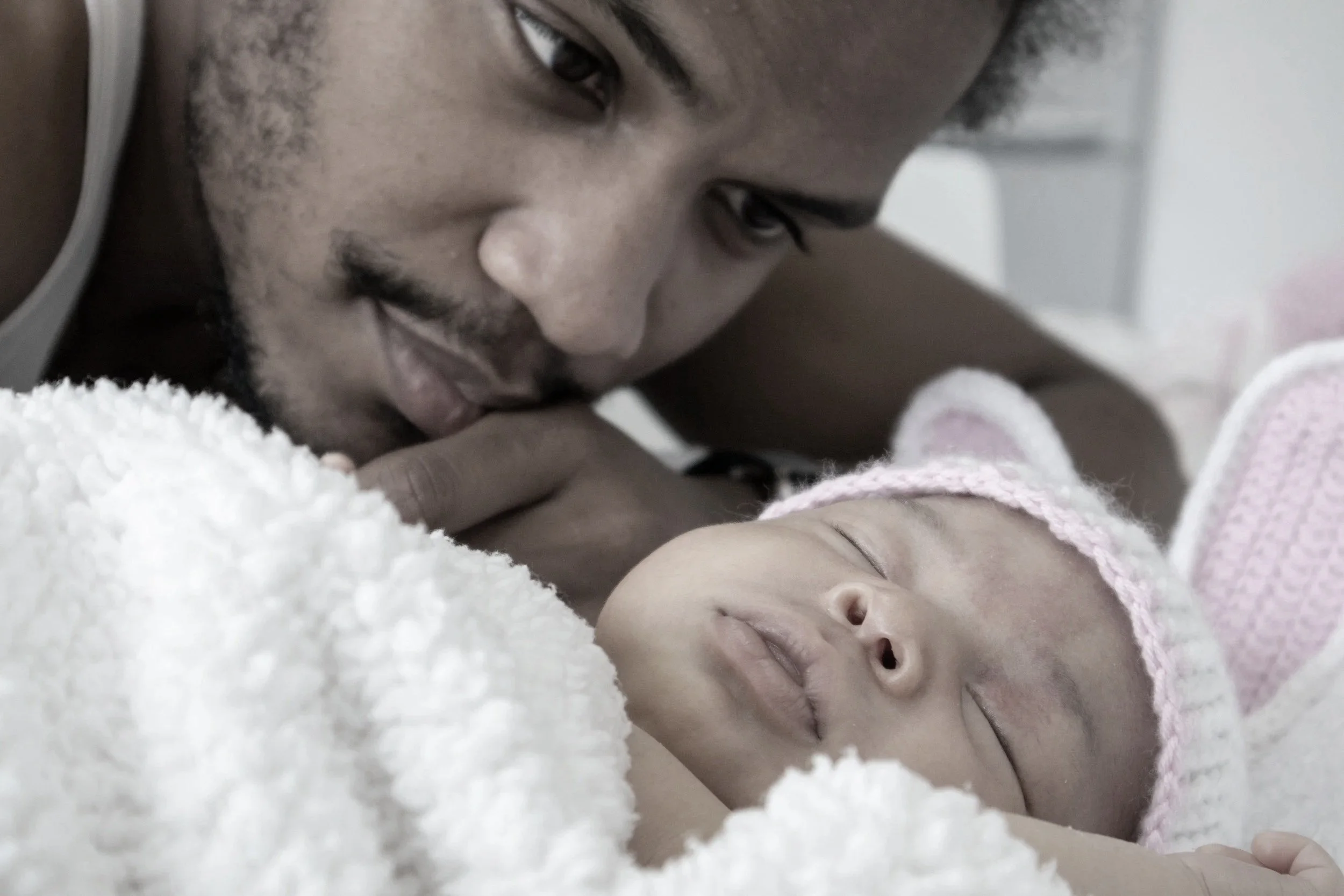 A man with short black hair and facial hair gently touches his forehead to a baby lying on a soft white blanket, both appearing calm and tender.