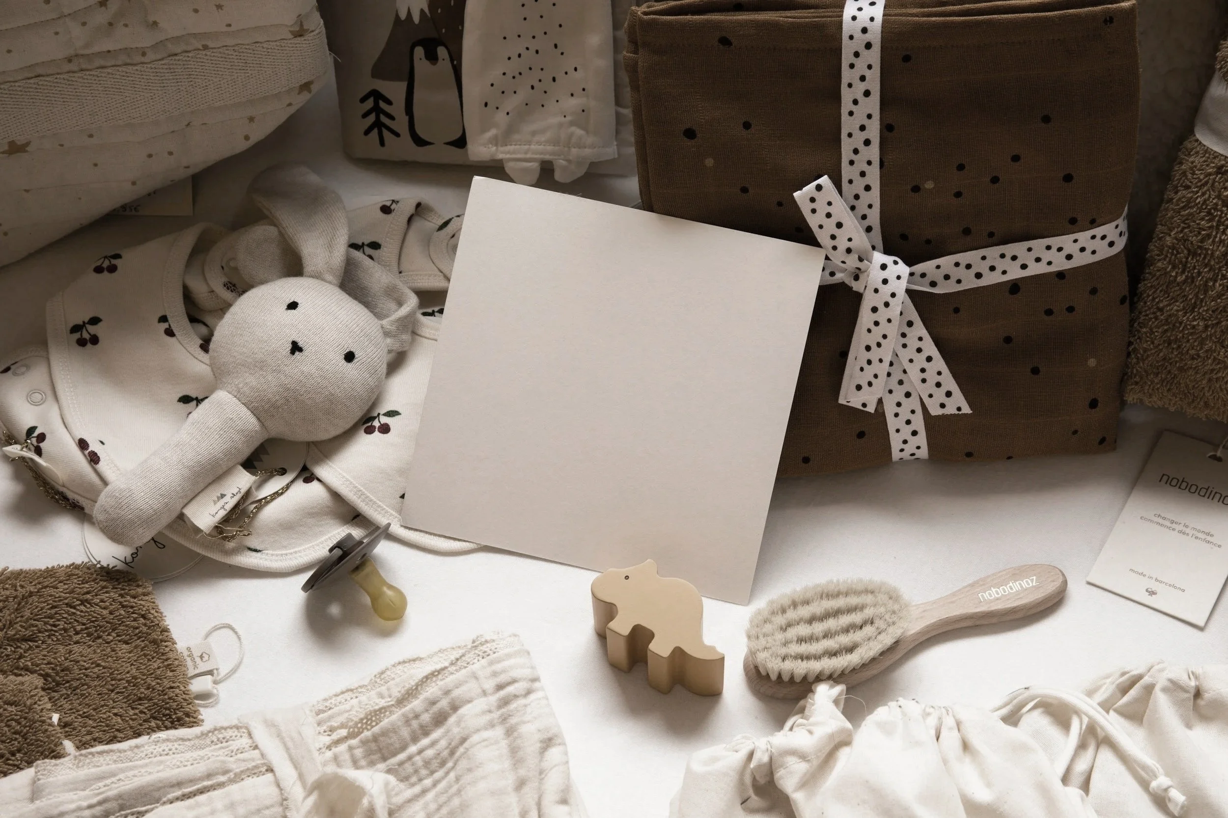Display of baby items including a brown gift box with polka dot ribbon, a pair of baby shoes, a plush bunny toy, and baby clothing and accessories in neutral and cream tones with small cherry prints and natural textures.