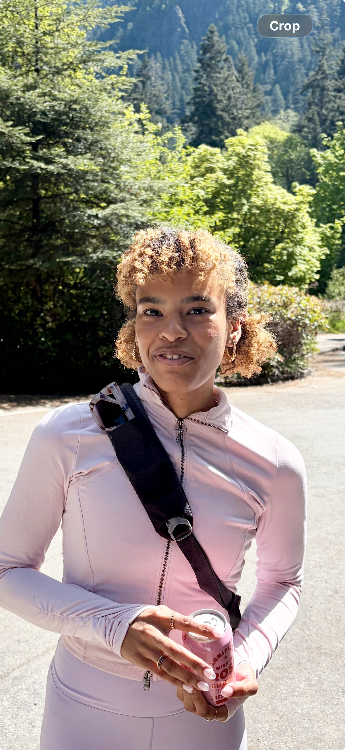 Young woman with curly blonde hair, earrings, and a light pink athletic jacket holding a drink, standing outdoors with lush green trees and mountains in the background.