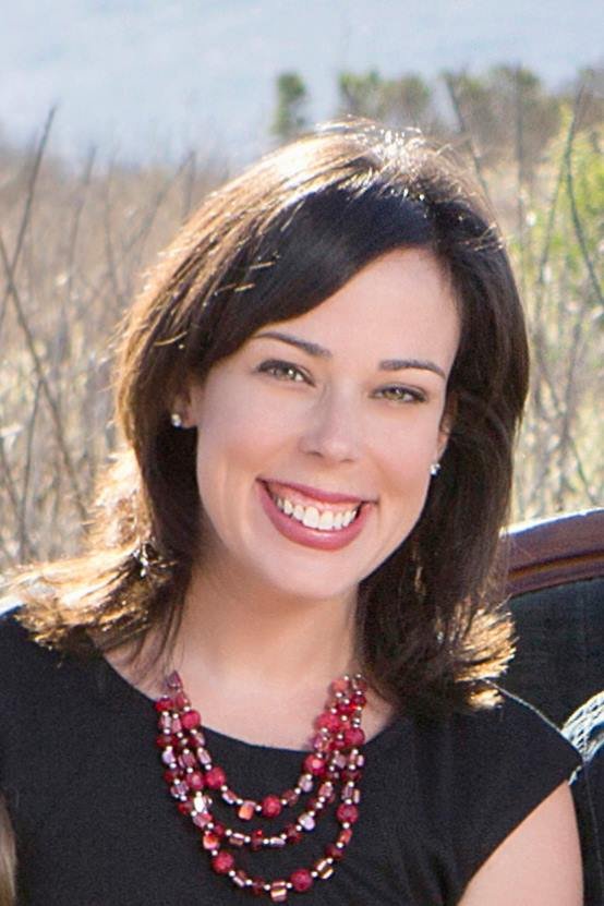 A woman with shoulder-length brown hair smiling outdoors, wearing a black top and a multi-strand red necklace.