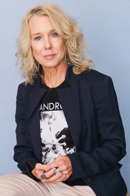 A woman with curly blonde hair wearing a black graphic T-shirt and a dark blazer, sitting against a light blue background.