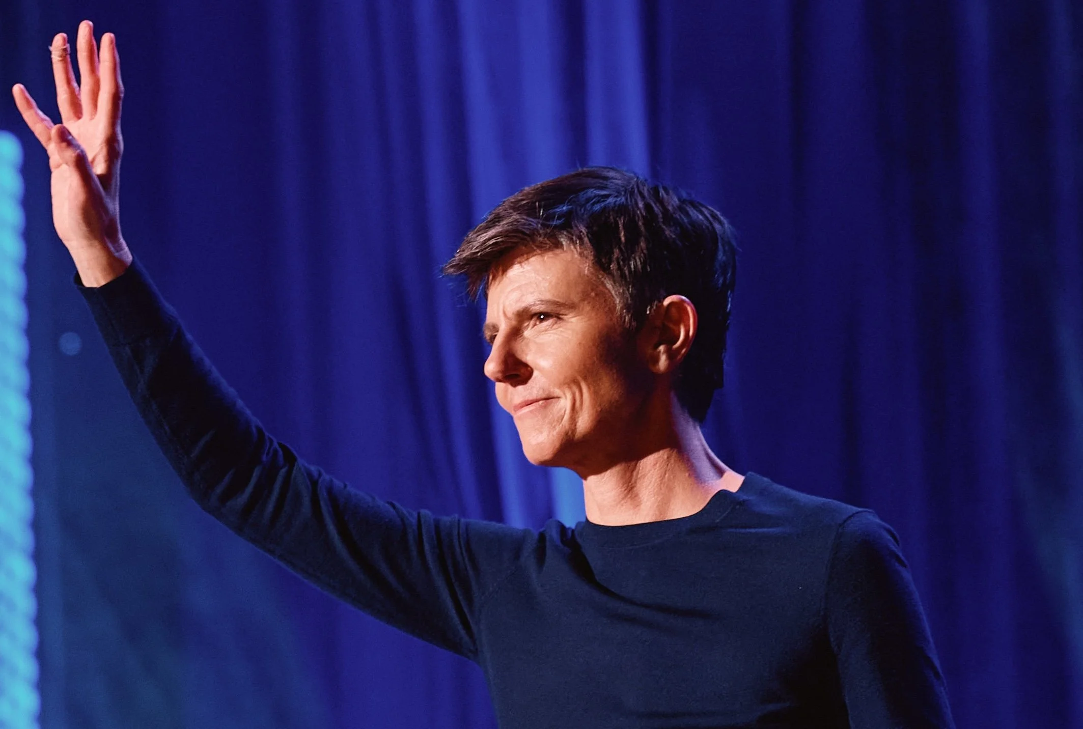 A woman with short dark hair and wearing a dark long-sleeve shirt is standing in front of a blue curtain, raising her right hand.