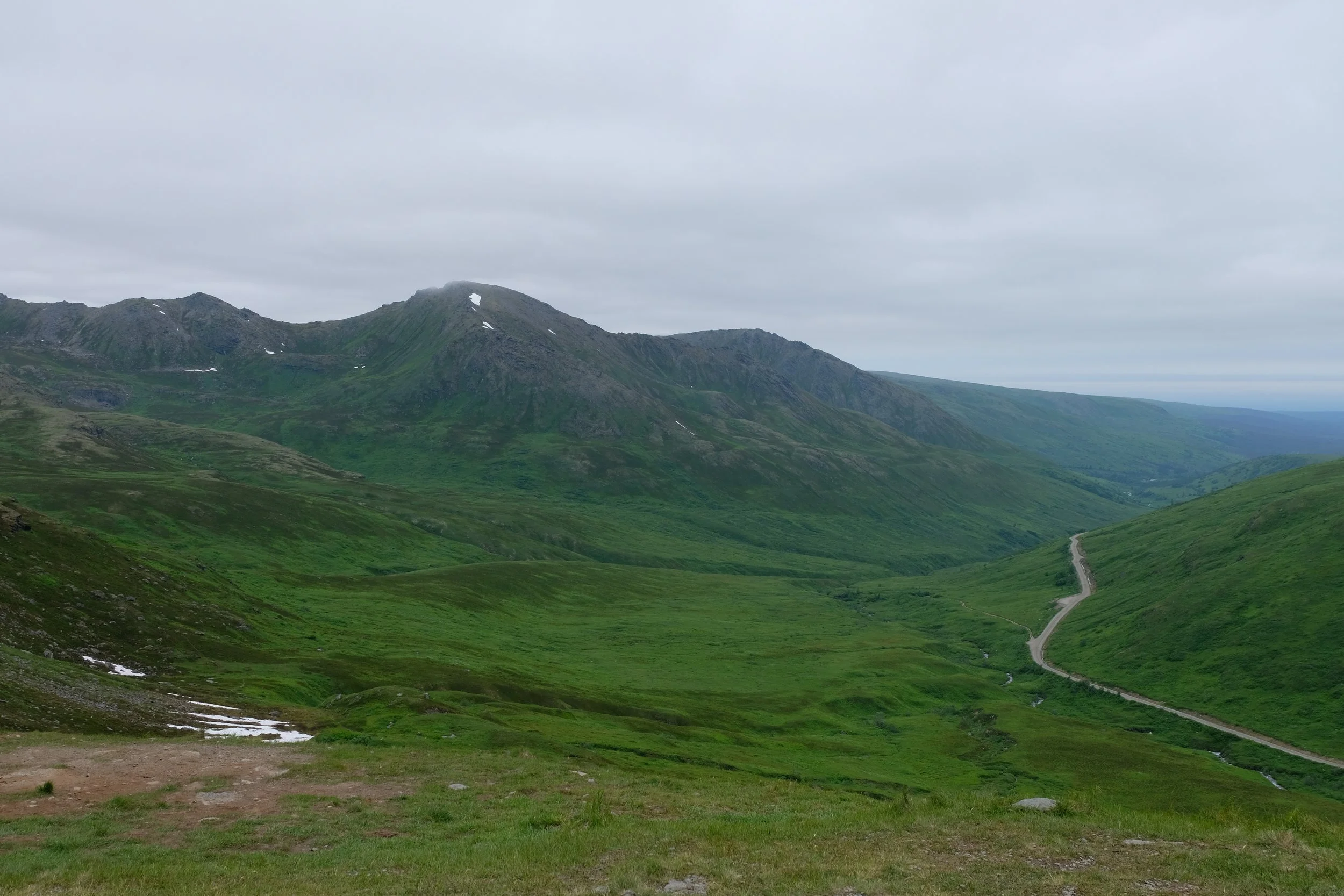 Green rolling hills with a winding road and mountains under a cloudy sky.