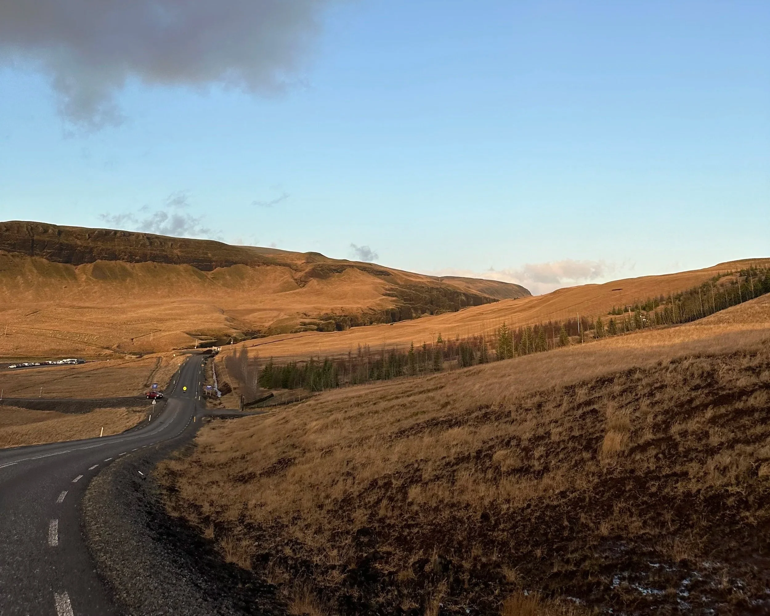 Scenic view of a winding road through a hilly countryside with dry grass and sparse trees under a partly cloudy sky.