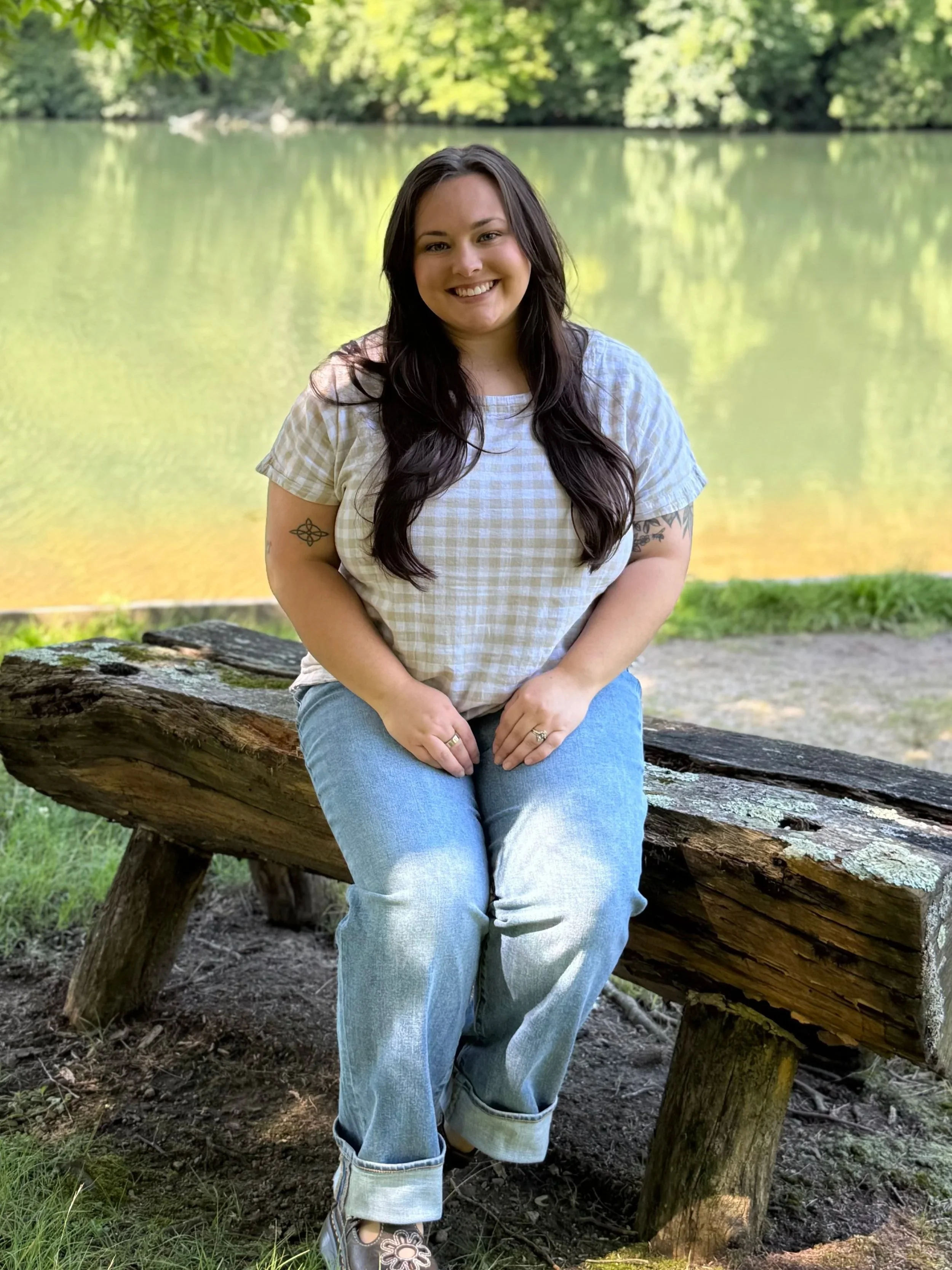 A smiling woman with long dark hair sitting on a large log near a lake, surrounded by green trees.