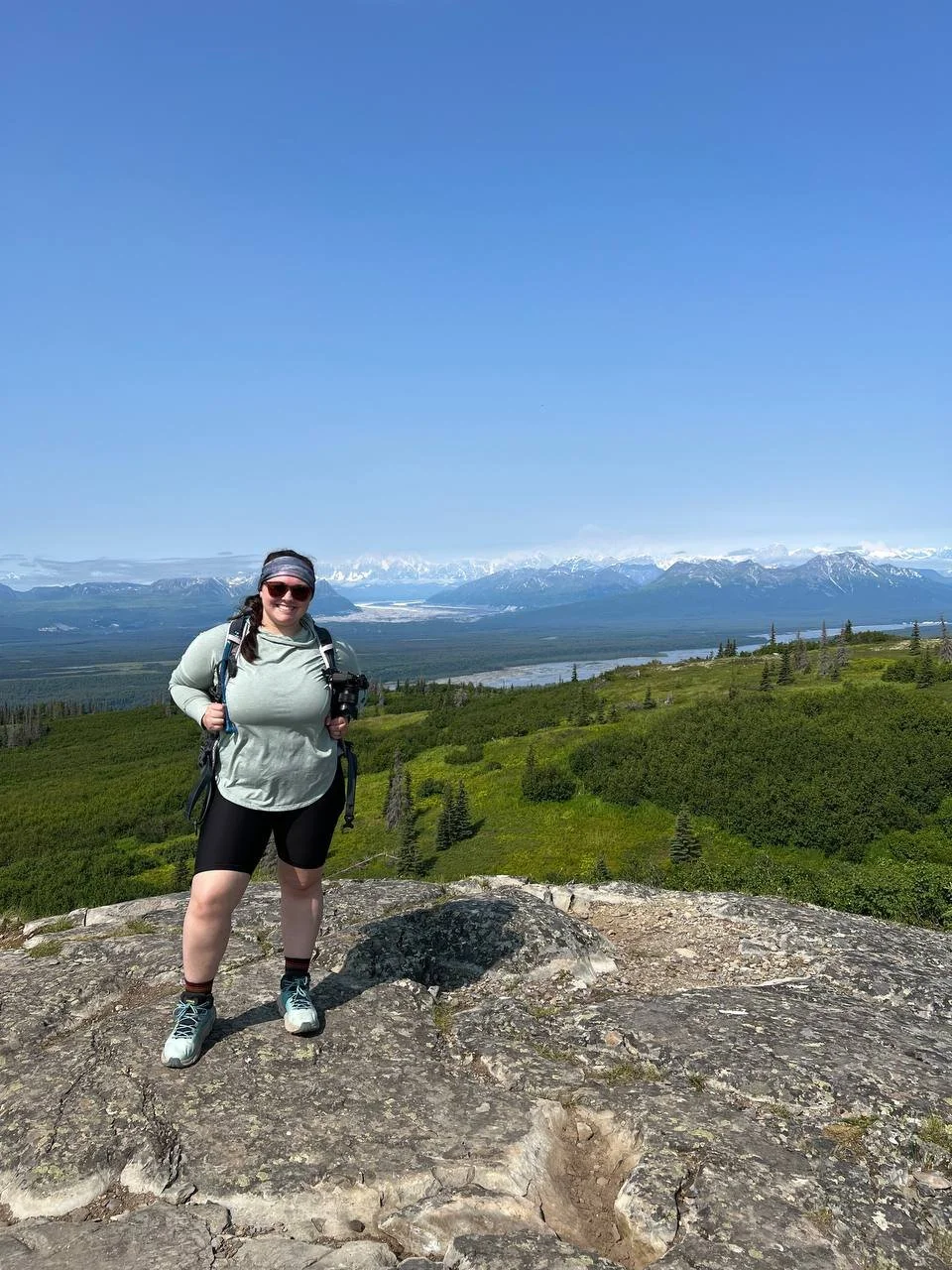 A woman in outdoor hiking gear standing on a rocky surface with a scenic view of green mountains, a river, and snow-capped peaks in the background under a clear blue sky.