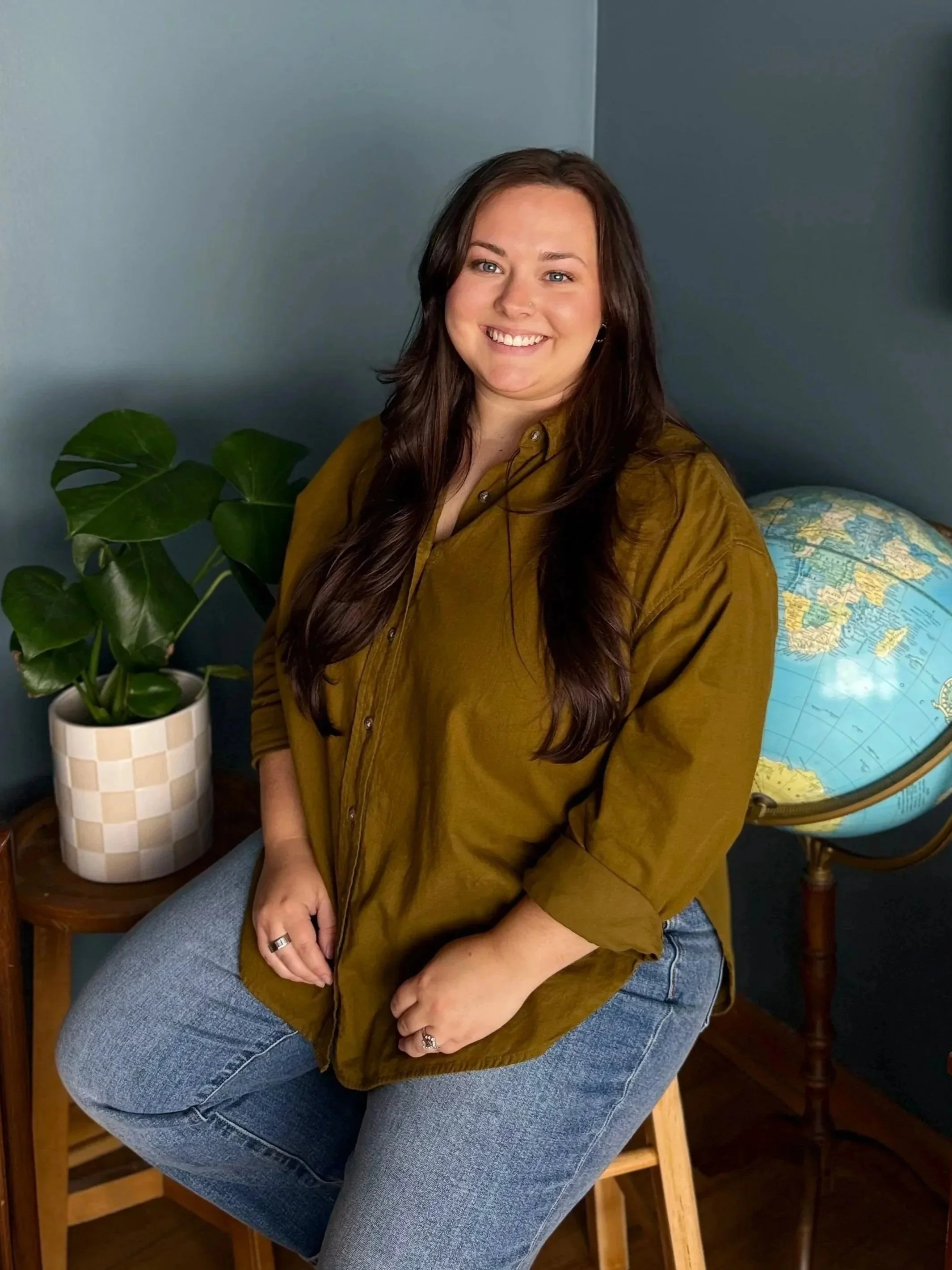 A woman smiling, wearing a mustard-colored shirt and blue jeans, sitting with her hands resting on her lap, beside a potted plant, in front of a globe on a dark background.