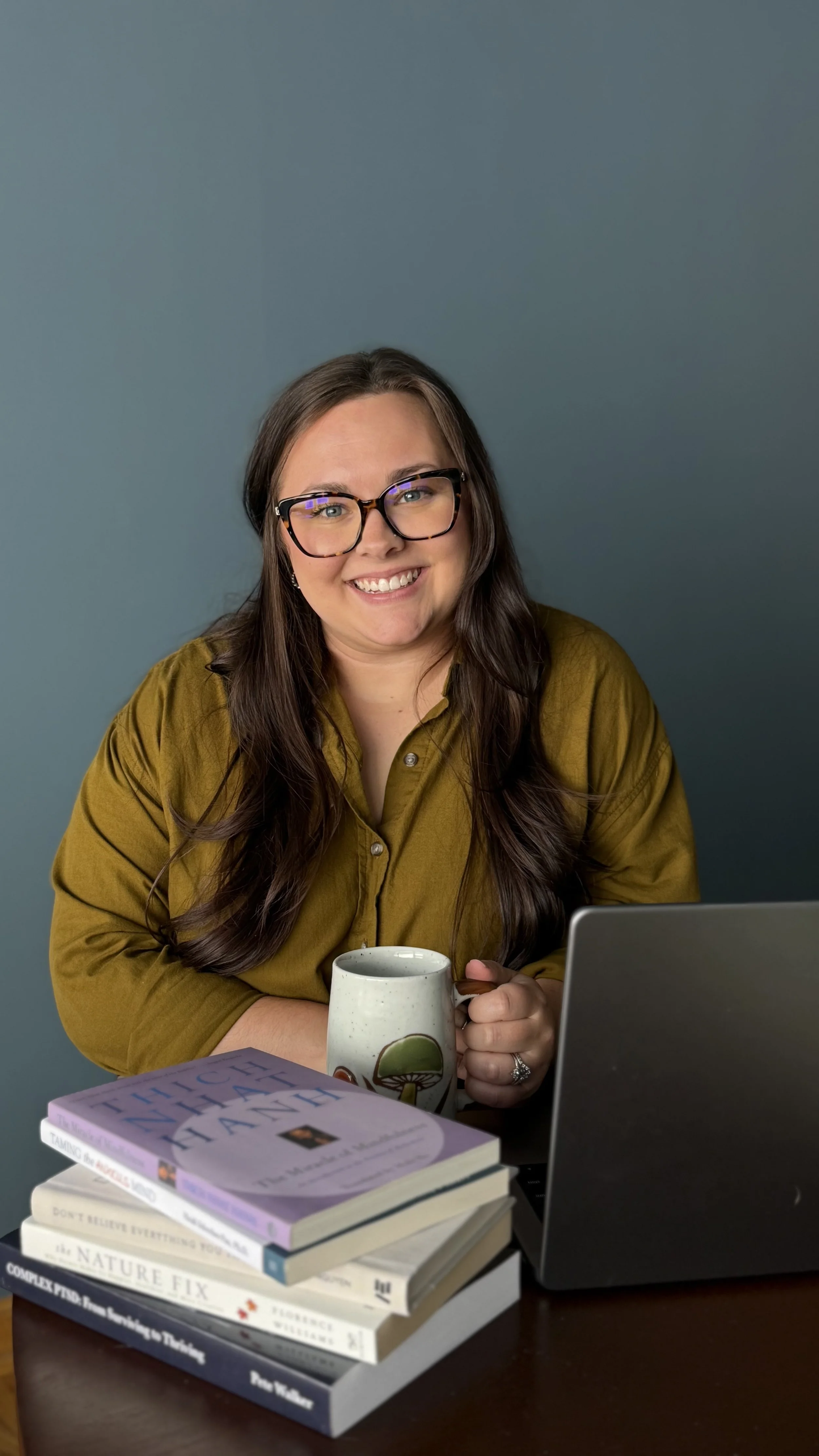 A woman with long brown hair, glasses, and a mustard-colored shirt smiling at the camera, sitting at a desk with a laptop, a stack of books, and holding a coffee mug.