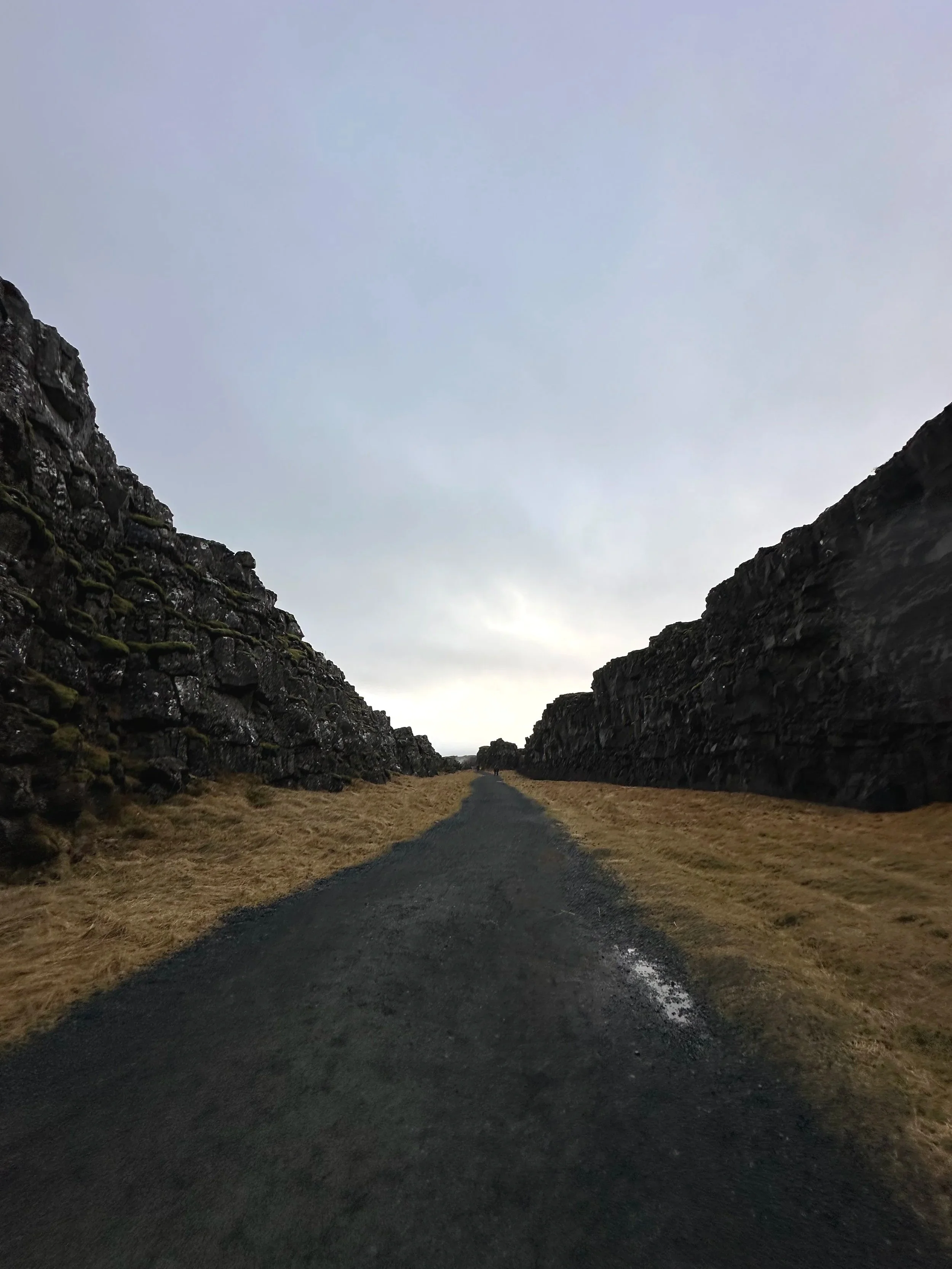 A narrow asphalt path runs between rocky cliffs with moss and grass, leading towards a cloudy sky at sunset.