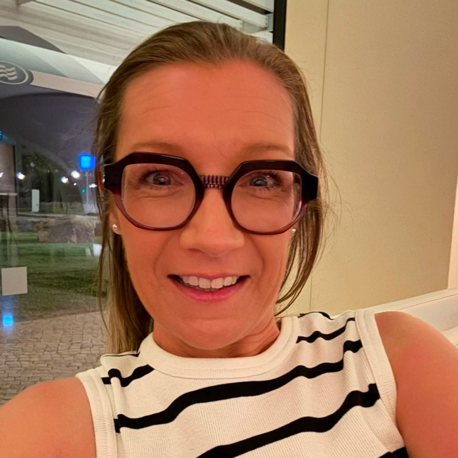 Sound Waves Choir Member - Sarah taking a selfie indoors, wearing glasses and a white sleeveless top with black stripes, with a window showing outdoor lights and landscape in the background.