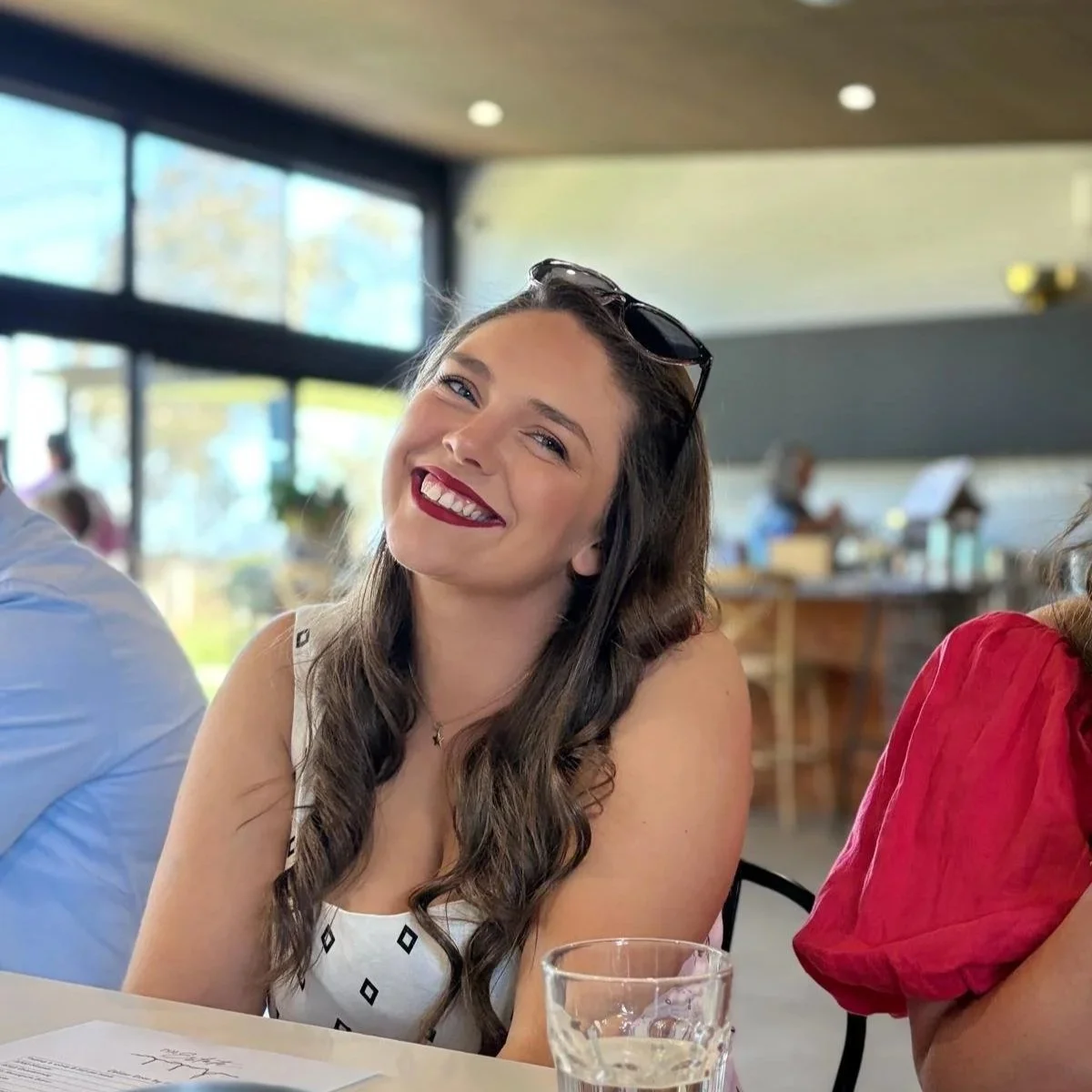 Sound Waves Member Kim wearing a white dress with black diamond patterns and sunglasses resting on her head, sitting at a table in a bright, modern cafe.