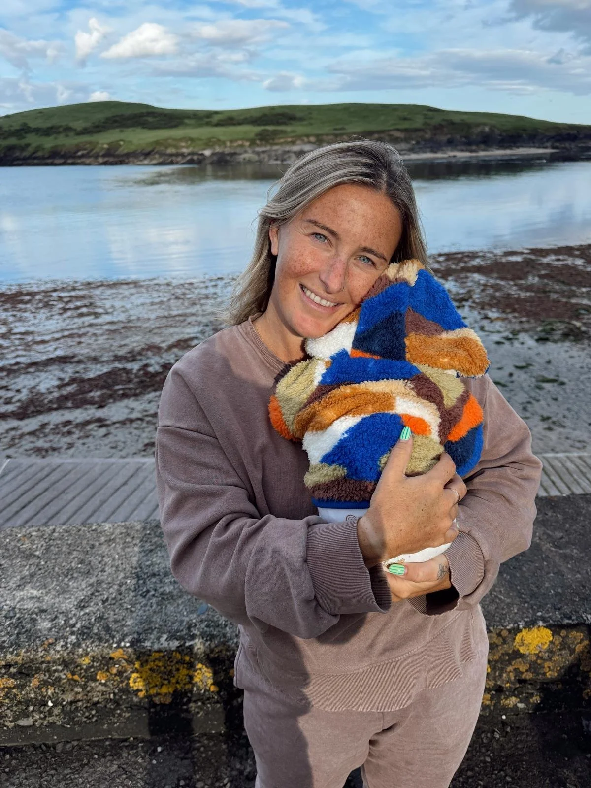 Sound Waves Member Jen stands smiling while holding a dog wrapped in a colourful, fluffy blanket. She's outdoors near a body of water with green hills and a cloudy sky in the background.