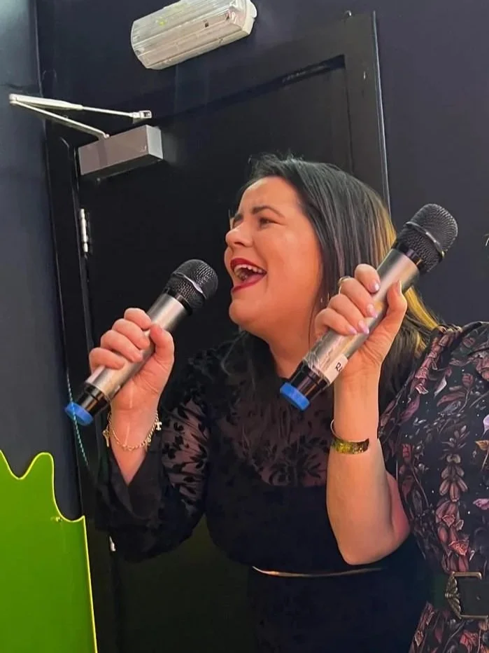 Sound Waves member Triona singing a microphone, smiling, with a dark background and part of a green object visible in the lower left.